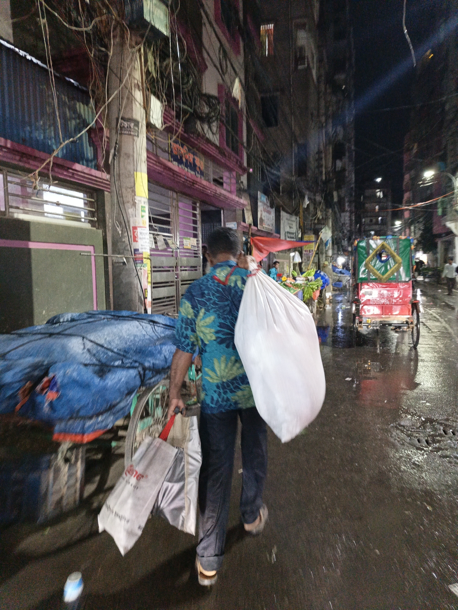 A man in a green and blue patterned shirt walks away from the viewer on a wet street, carrying a large white bag over his shoulder and silver shopping bags in his hand. The location appears to be in Dhaka, Bangladesh, based on the presence of a cycle rickshaw in the background and the architectural style of the buildings. The man is walking past a covered cart or stall on the left. A brightly colored cycle rickshaw is visible further down the street, with other vendors and pedestrians. The street is slick with water, reflecting light from street lamps and shopfronts. Buildings line both sides of the street, with dense overhead wires. It is nighttime.