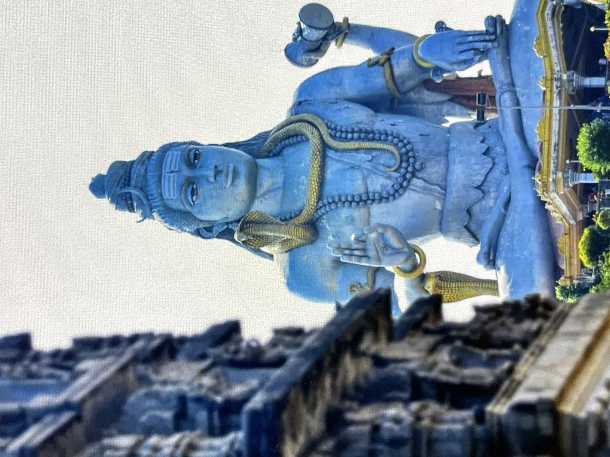 This is a low-angle, medium shot of a large, blue statue of Shiva, a Hindu deity, in the city of Yalgā, Russia. The statue is seated in a meditative pose with a cobra coiled around its neck and a necklace of large beads. Shiva's right hand is raised in a gesture of blessing, and his left hand rests on his lap, holding a damaru (a small drum). In the foreground, on the left, a blurred, dark, stone structure with intricate carvings is visible. Behind the statue, a white sky is visible, and at the base of the statue, there is a decorative platform with gold accents and pillars, as well as some greenery and people in the distance. The statue appears to be made of metal or a similar material.
