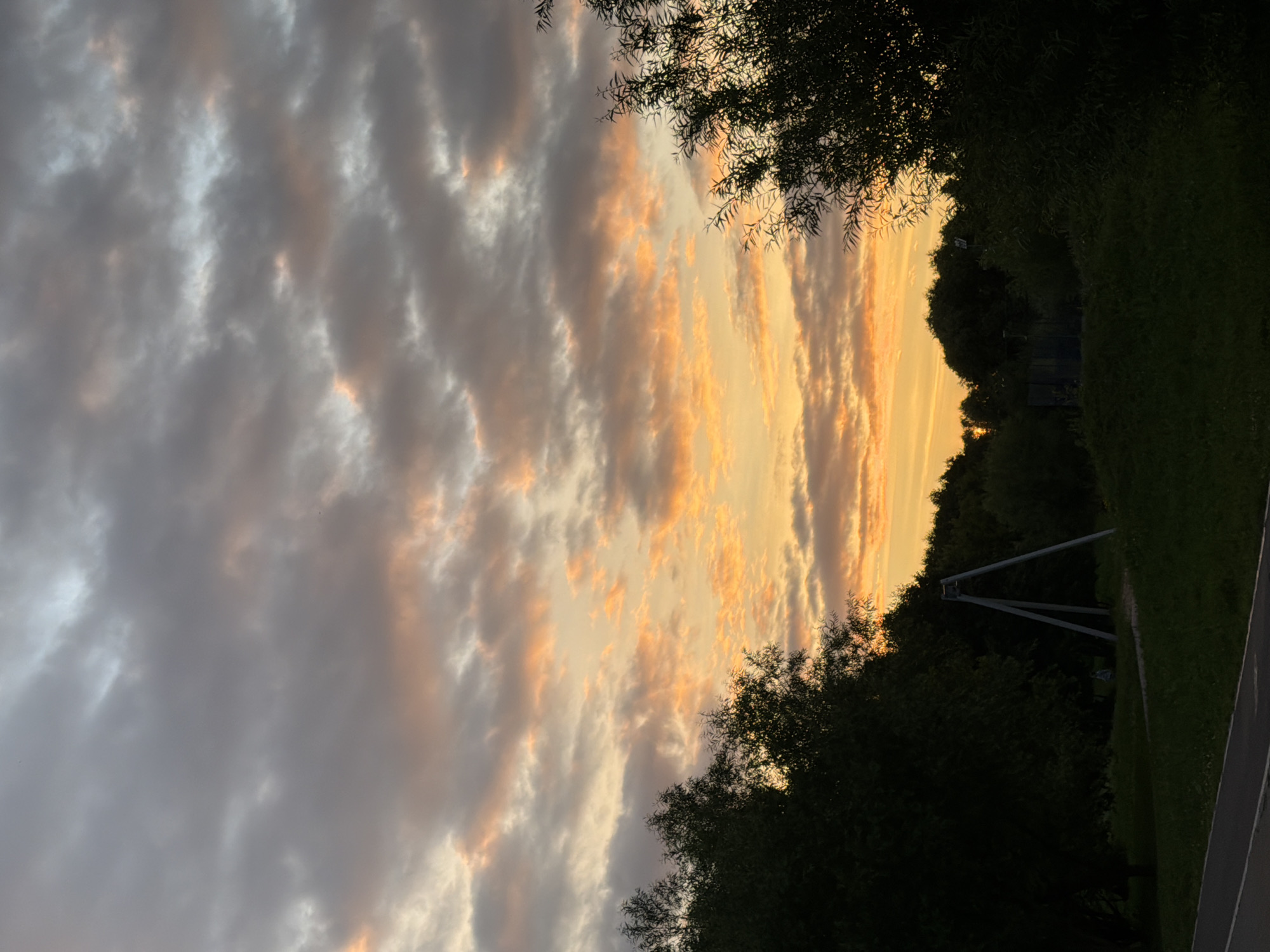 The image shows a sunset over a grassy area with trees. The sky is filled with clouds in shades of grey, white, and orange, indicating a dramatic sunset. In the foreground, dark green trees frame the scene on the left and right. A light-colored path is visible on the right side of the image. In the background, there are more trees and a metallic structure, possibly part of a sports facility or playground equipment. The lighting suggests it is late evening, as the sun is setting. There is no visible text in the image. The overall mood is peaceful and serene, capturing the beauty of a natural sunset.