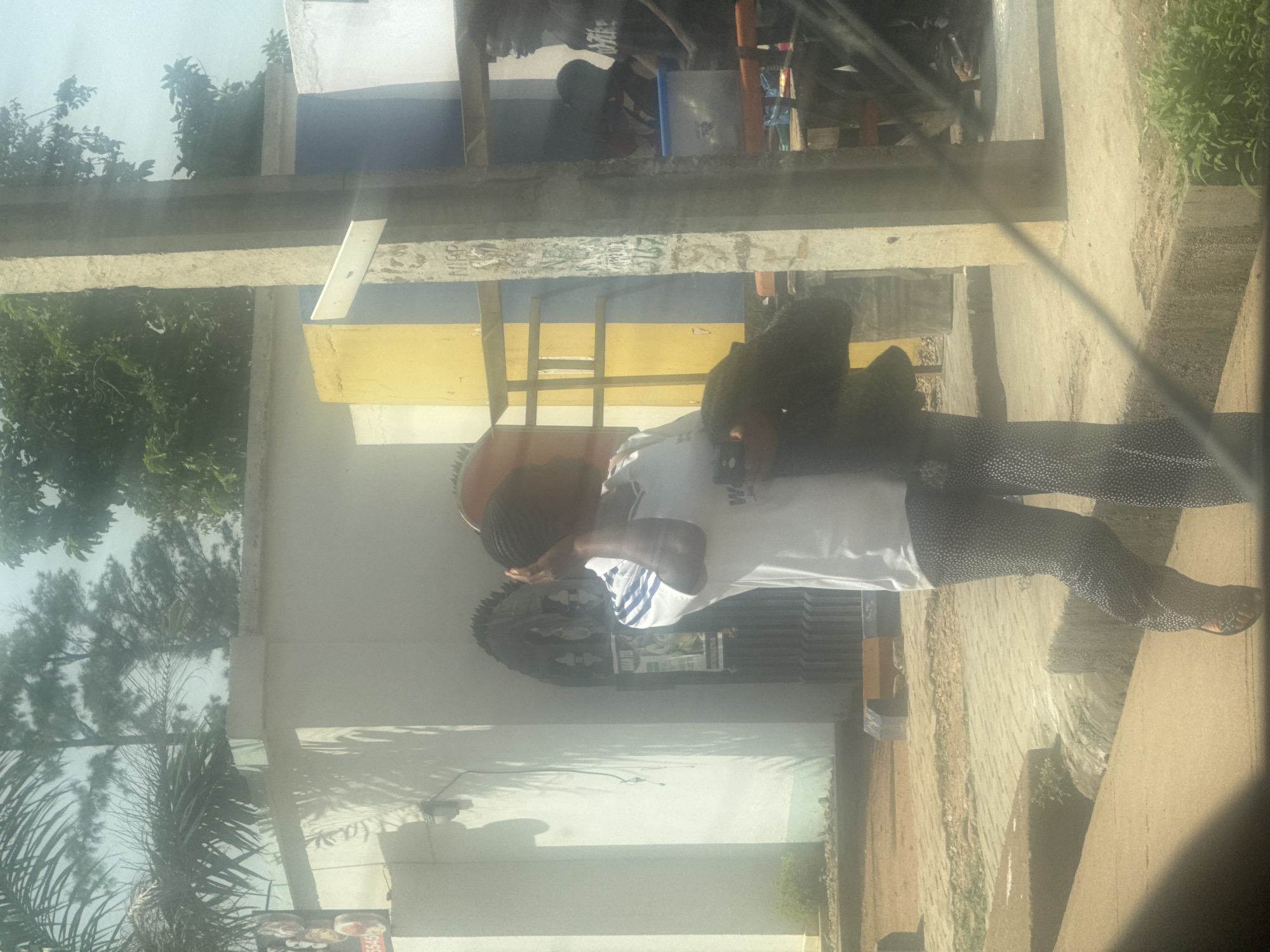 The image shows a person in the foreground, facing away from the camera and towards the right. They are wearing a white t-shirt with blue stripes on the sleeves and black pants with white polka dots. The person appears to be interacting with something held in their hands, which are partially obscured. Behind this person, there is a building with a white facade and a yellow and blue section. There are also trees and greenery visible, suggesting an outdoor setting. The scene is lit by daylight, though the exact time of day is difficult to ascertain due to the angle and quality of the image. The image appears to have been taken from a vehicle, indicated by a reflection on the left side of the frame. The overall impression is that of a casual outdoor scene in a community area. The image is oriented vertically, with the top of the frame at the bottom.
There are no clear indications of the weather or specific emotions conveyed by the visible subjects. Location cues are limited to the building and greenery, but without more information, it's hard to pinpoint a precise location. There is some faint text on the building that is illegible.