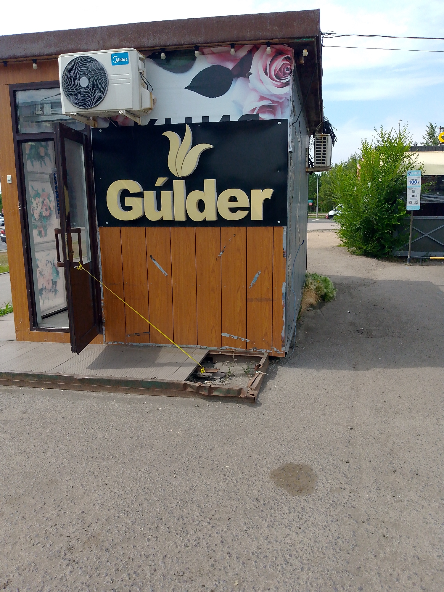 The image captures a street-level view of a small commercial kiosk in Astana, Kazakhstan. The kiosk features a dark signboard with a golden tulip logo and the word "Gúlder" written in large, golden, serif font. Above the signboard, a large graphic of pink roses is visible. An outdoor air conditioning unit, branded "Midea," is mounted on the wall above the entrance. The entrance itself is a brown wooden door, ajar and secured by a yellow string tethered to the ground. The kiosk is situated on a weathered wooden platform that extends onto the asphalt road. Several metal frames and debris are present on the ground around the kiosk, suggesting a state of disrepair or ongoing construction. To the right of the kiosk, a green leafy bush grows, and further in the background, a white vehicle is parked. A blue sign with "100₸" and a QR code is posted on a metal pole near the bush. The sky above is partly cloudy.