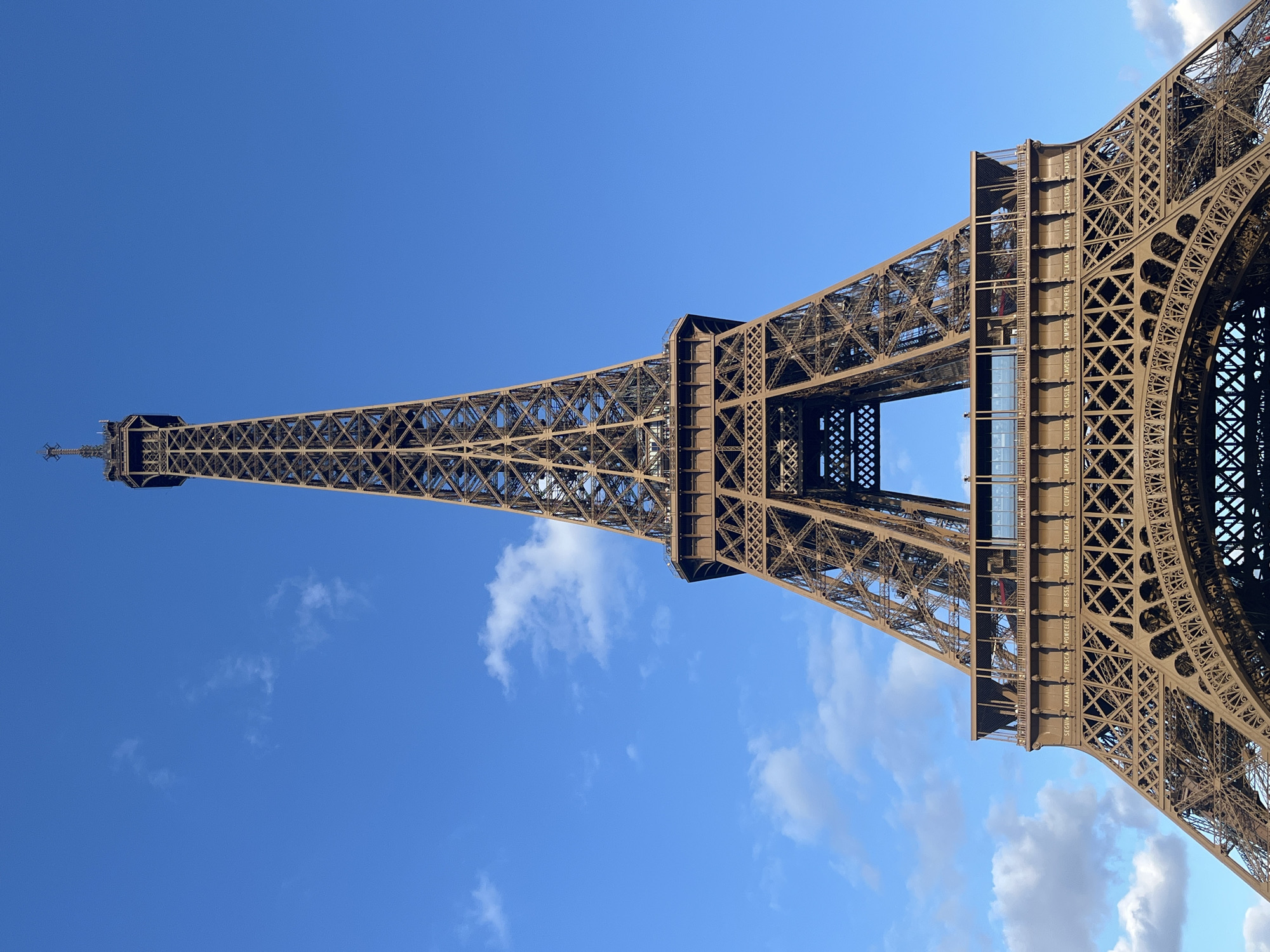 This media file shows an upward-angled, close-up view of the Eiffel Tower against a clear blue sky with scattered white clouds. The iron lattice structure of the tower dominates the frame, appearing in a warm, bronze color. The perspective emphasizes the immense scale and intricate design of the landmark.
The lighting suggests it is daytime, likely late morning or early afternoon, given the brightness of the sky and the shadows cast on the tower's structure. The weather appears pleasant and sunny, with only a few wispy clouds adding texture to the sky. There are no people visible in the frame, focusing solely on the architectural marvel.
Several lines of text are visible inscribed vertically on the beams of the tower, most prominently on the lower sections of the structure. These appear to be names of scientists and engineers, a common feature on the Eiffel Tower. The visible text reads: "SEGUIN LALANDE TRESCA PONCELE BRESSE LAGRANGE BELANGE CUVIER LAPLACE DULONG CHASLES LAVOISIEM AMPERE CHEVREU FLACHAT NAVIER LEGENDRE CHAPTAL". The image is oriented with the tower extending upwards from the bottom right to the top left of the frame.