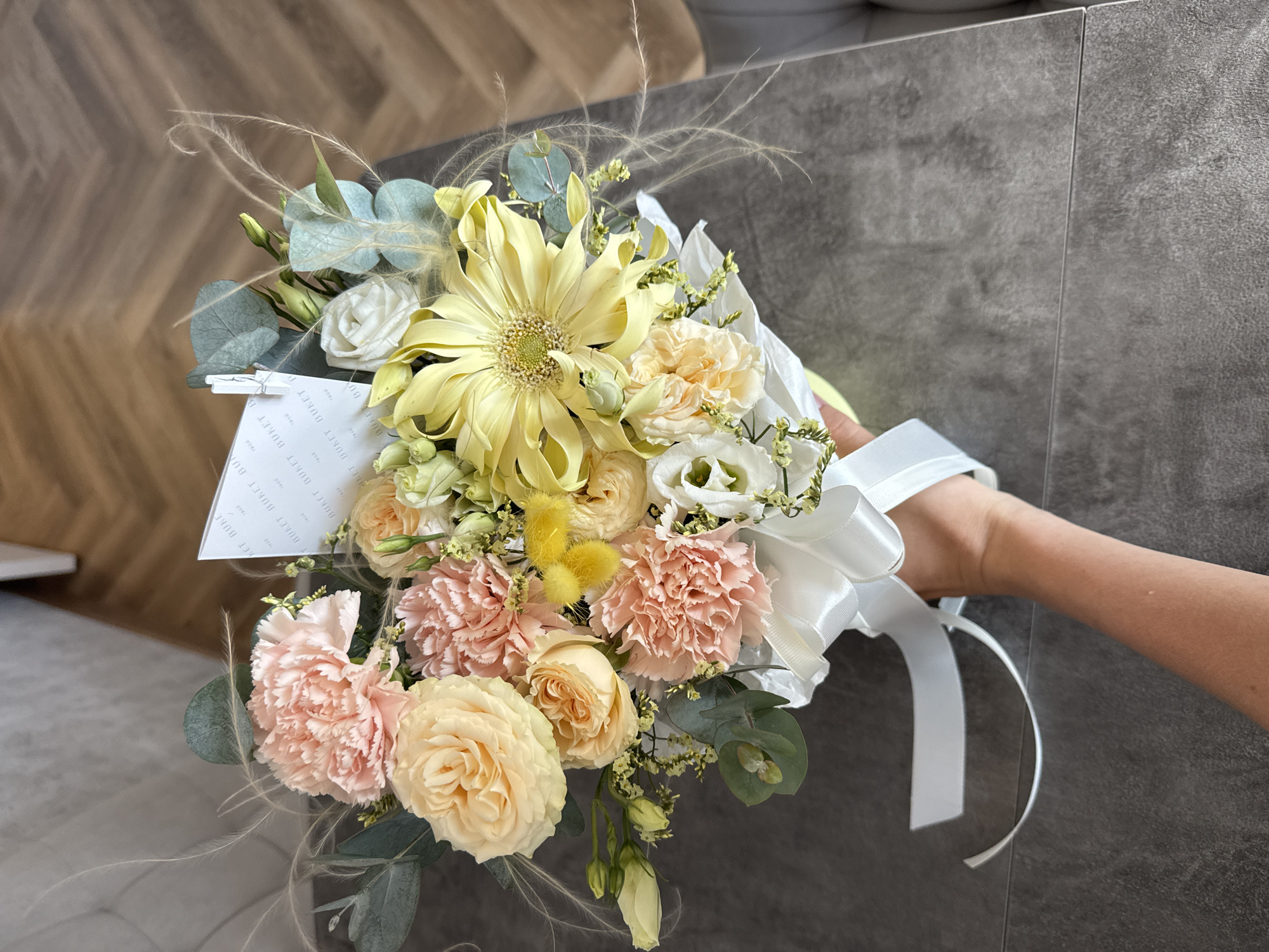 A hand holds a bouquet of flowers with a white ribbon tied around it. The bouquet includes yellow and peach-colored roses, peach-colored carnations, white lisianthus, yellow billy buttons, and eucalyptus leaves. The flowers are wrapped in white paper. The hand is in the lower right of the image. The background is a gray wall on the right side of the image and a light brown wooden surface on the left. A small card with printed text is visible within the bouquet.