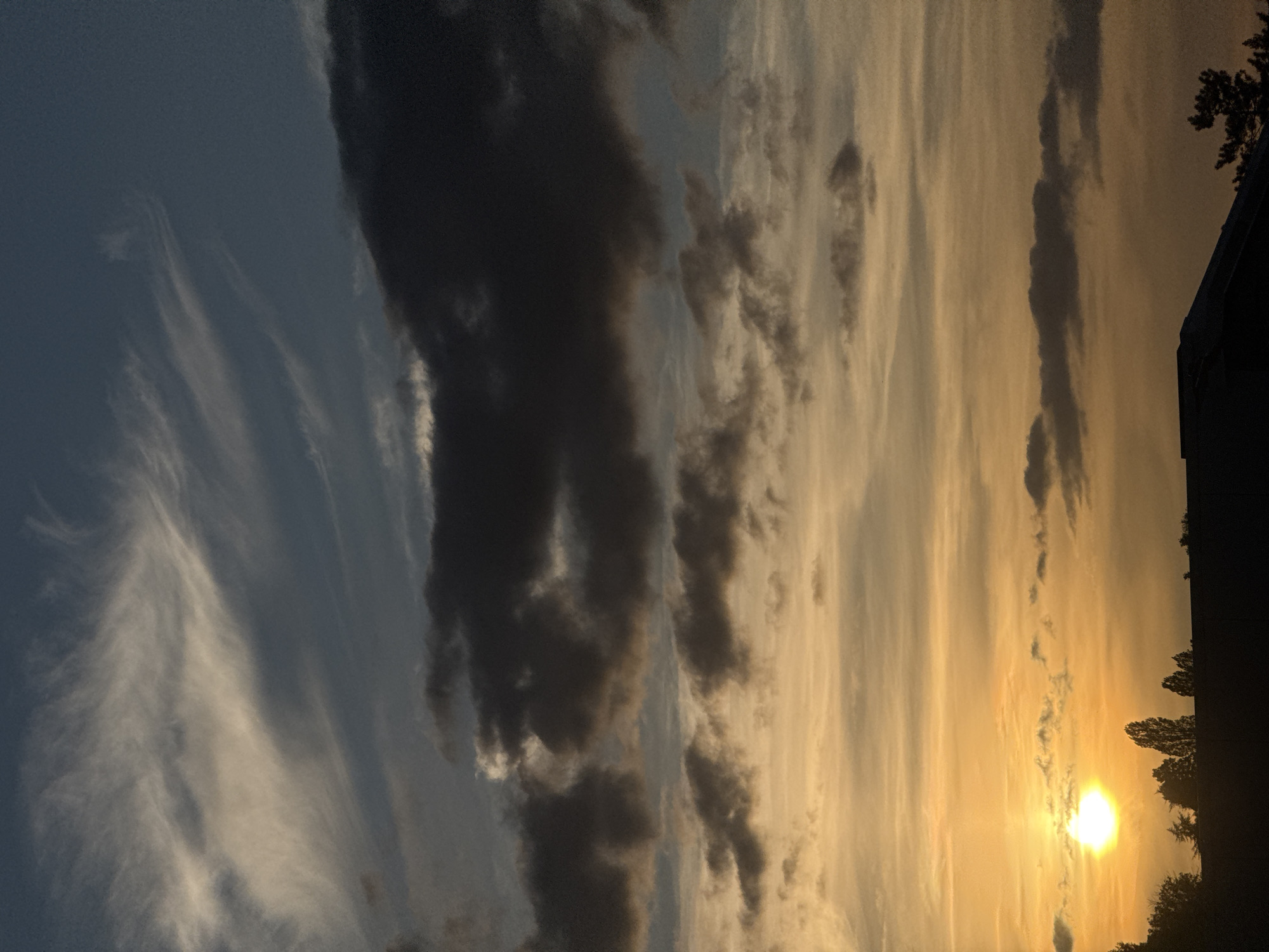 The image displays a dramatic sky scene captured during either sunrise or sunset in Bures-sur-Yvette, France. The sky exhibits a striking color gradient, transitioning from deep blue and dark grey on the left to a brilliant golden-orange hue on the right, where the sun is positioned.
A bright, intensely glowing sun disk is prominently visible in the lower right portion of the frame, partially diffused by light clouds, and casting a warm, golden light across the landscape. The sky is filled with a variety of clouds: delicate, wispy white cirrus-like clouds are visible against the darker blue on the left, while large, dense, and dark grey clouds dominate the central and right areas. These heavier clouds create strong contrasts, appearing almost black against the brighter sections of the sky and interspersed with streaks of lighter, golden-illuminated clouds.
In the far lower right corner, silhouetted against the vivid sky, are the dark forms of several trees, appearing dense and possibly evergreen. Adjacent to the trees, a dark, indistinct architectural structure, likely part of a building, is also visible as a silhouette, forming a clear horizon line. The scene conveys a sense of natural beauty and atmospheric depth, characteristic of a transitional period of day. No text is visible in the image.
