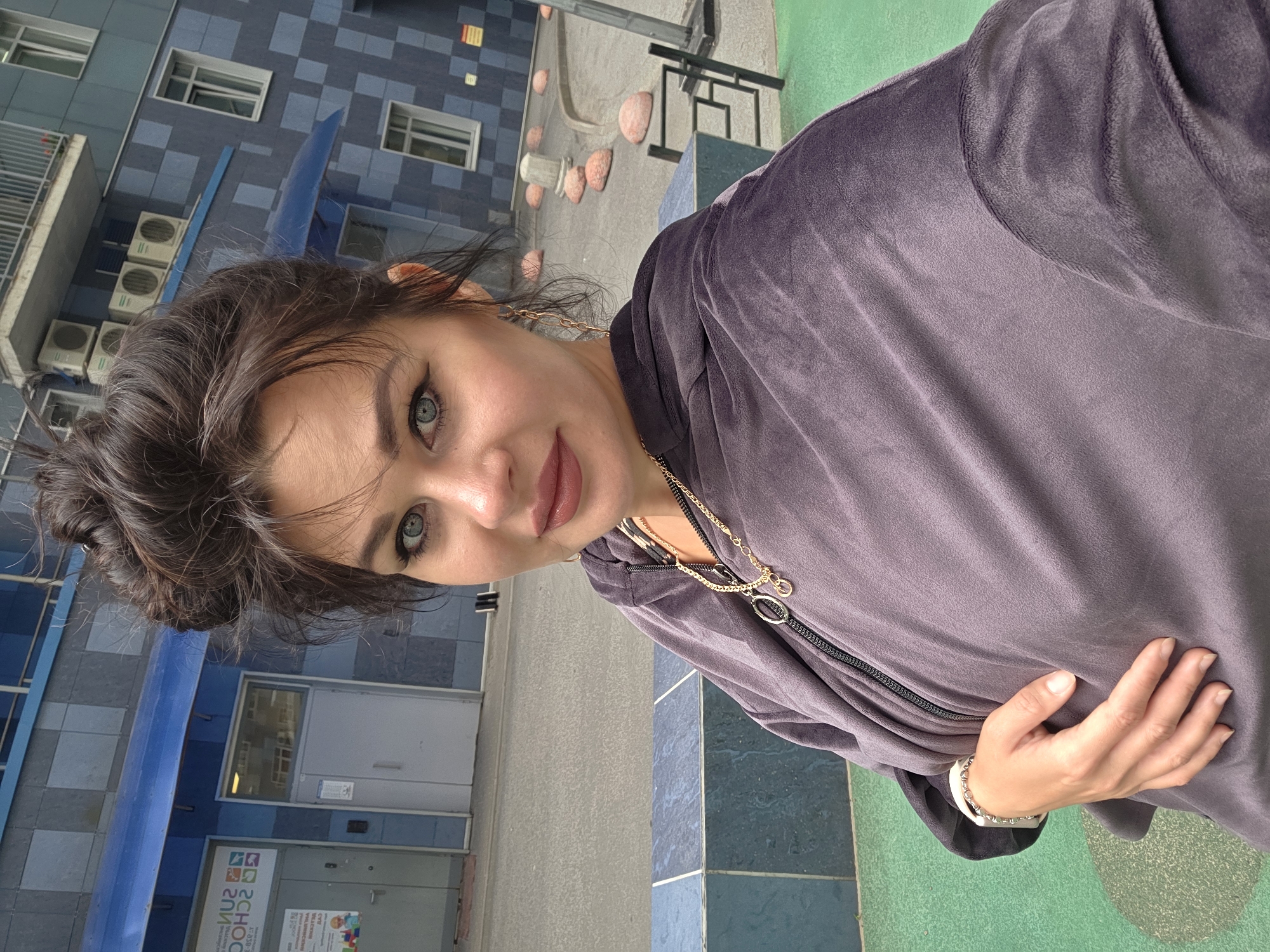 A woman with dark brown hair styled in a messy bun takes a selfie outdoors. She is wearing a purple velour hoodie and a silver bracelet. She has blue eyes and is wearing makeup, including eyeliner and lipstick. In the background, there is a blue building with several air conditioning units visible on its facade. The building appears to be a school or educational institution, with signage that includes the words "SUN SCHOOL." A paved area with some round, reddish stones is also visible. The ground in the foreground is green. The overall scene suggests an urban environment, possibly a schoolyard or a courtyard in Saint Petersburg, Russia.