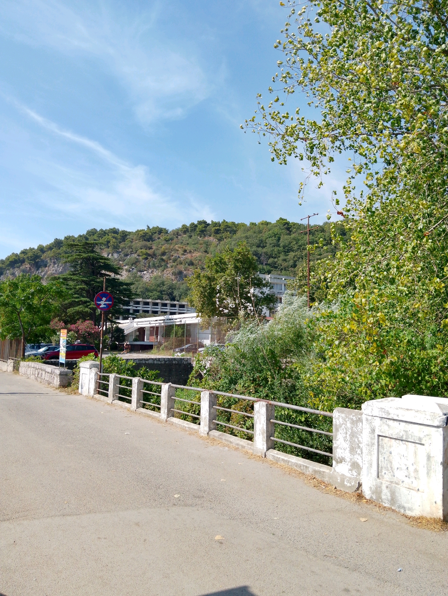 The image depicts a sunny day with a clear blue sky and scattered white clouds. In the foreground, an asphalt road runs alongside a bridge with a concrete railing. The bridge has metal bars supporting the railing and concrete posts at intervals. Beyond the bridge, a lush green hillside rises, dotted with trees and some rocky outcrops. In the middle ground, there is a low stone wall with vegetation growing behind it. A building with a white facade and a flat roof is visible behind the vegetation. Several cars are parked in front of the building, including a red vehicle. A traffic sign with a red circle and a horizontal bar indicating a prohibition is also visible. The overall scene suggests a location in a coastal or mountainous region. Given the context, this is likely a view of a street or road in Sutomore, Montenegro.