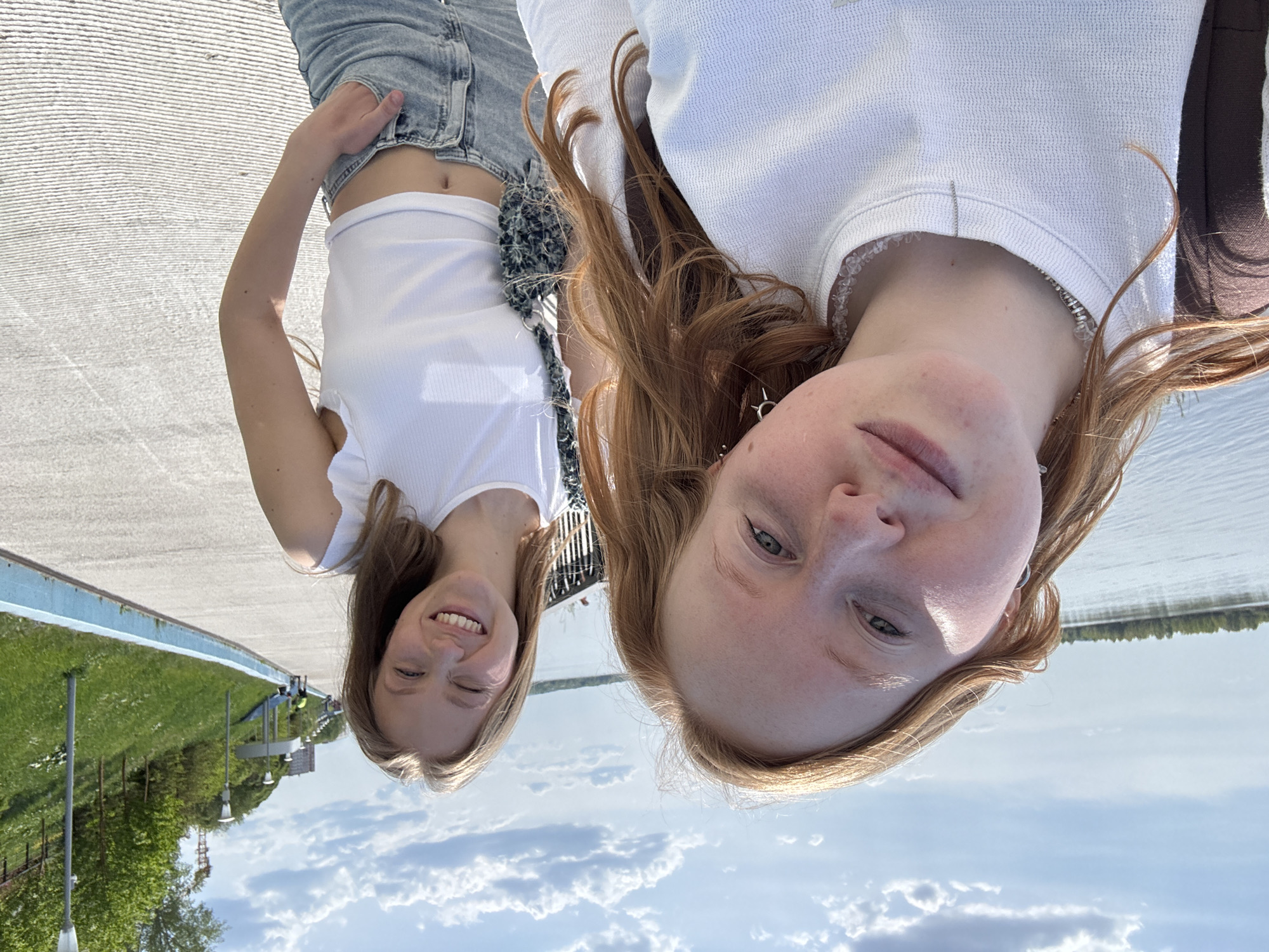 Two young women are posing for a selfie outdoors. The woman on the left is blonde, wearing a white crop top and blue jeans. She is smiling. The woman on the right has red hair and is wearing a white top with a beaded neckline. The background shows a blue sky with clouds, water, and green landscape. The location is likely a park or riverside area in the city of Perm, Russia.