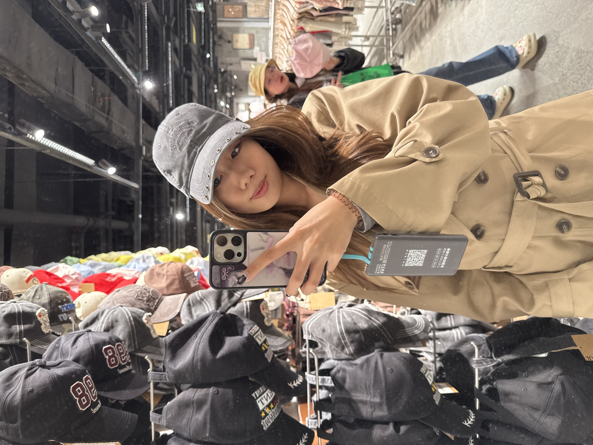 A woman in a gray cap and beige trench coat takes a selfie with an iPhone in a store, surrounded by stacks of baseball caps on metal racks. Another person in a straw hat and pink hat is visible in the background near a clothing rack. The location is the Jianlan Road District, China.