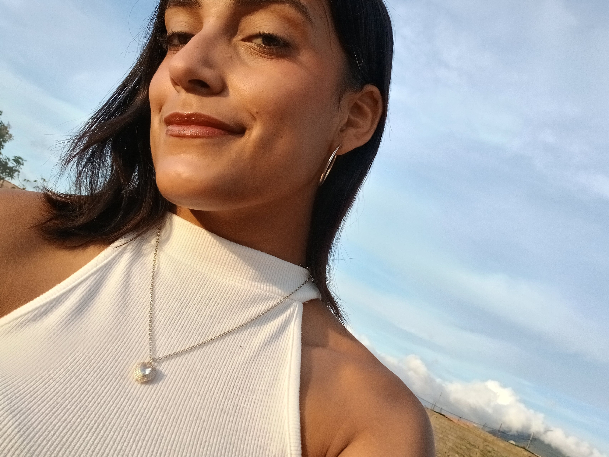 A close-up, half-body outdoor portrait features a young woman in Altagracia, Venezuela, captured during the golden hour of a late afternoon or early evening. She is positioned on the left side of the frame, looking directly at the camera with a subtle, pleasant smile and her head tilted slightly. Her dark, shoulder-length hair is parted on the side and appears slightly windswept.
She is wearing a white, ribbed sleeveless top and accessorizes with large silver hoop earrings and a silver chain necklace featuring a heart-shaped pendant. The warm, soft light illuminates the right side of her face and shoulder, highlighting her features and adding a glowing tone to her skin and hair. Her lips have a glossy finish.
The background primarily consists of a clear, light blue sky with scattered white clouds. To the right and lower part of the background, a gentle slope or grassy hill can be discerned, with hints of clouds resting closer to the horizon and possibly some distant structures or a fence. The outdoor setting and the warm lighting suggest pleasant weather. The woman appears calm and content. There is no visible text in the image.