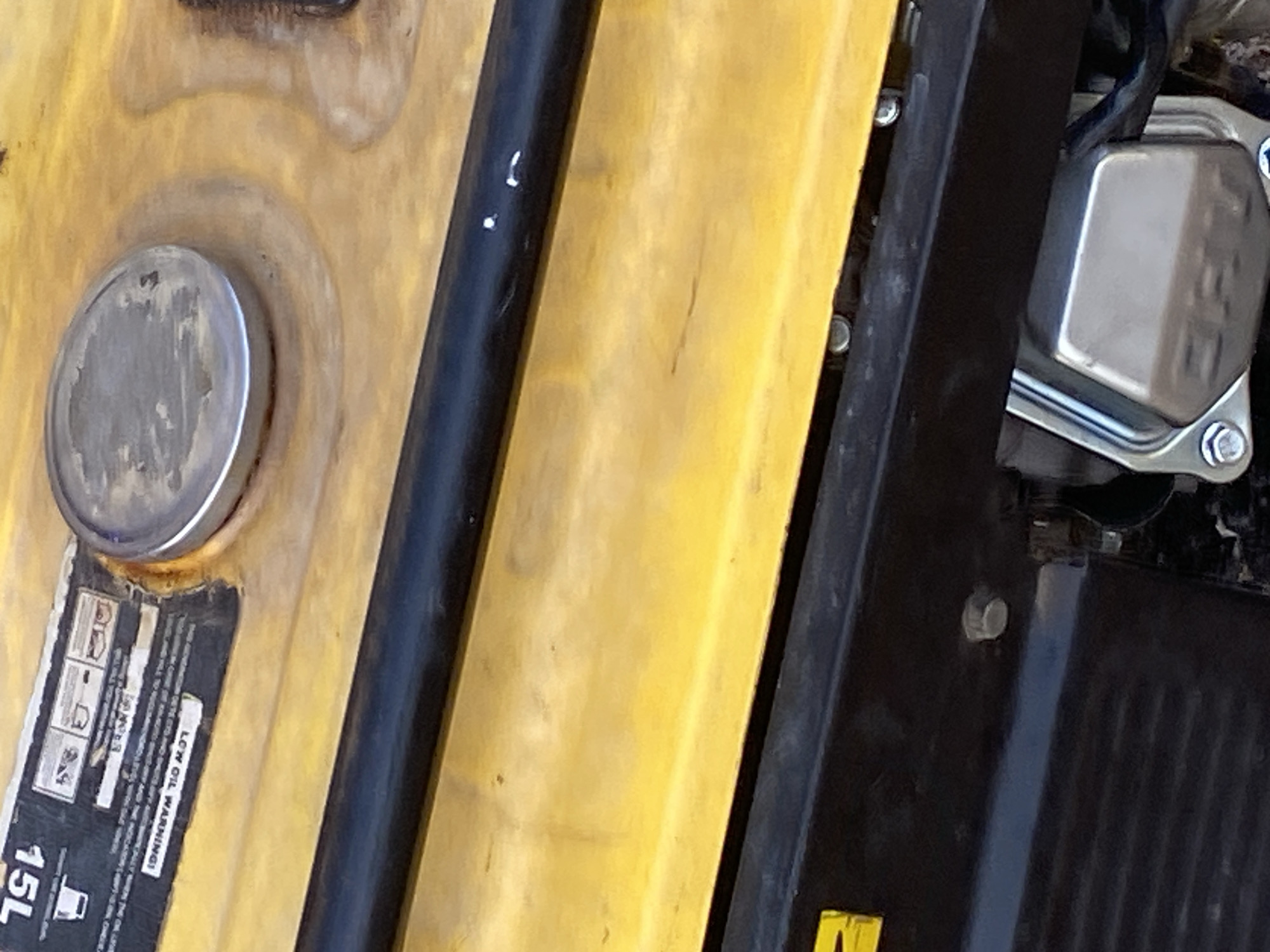 This image shows a close-up, overhead view of a yellow generator. A black metal rod runs horizontally across the upper portion of the frame, obscuring some of the generator's surface. To the left of the rod, a label with white and black text is visible, reading "15L" at the top and "LOW OIL WARNING!" further down. A silver, circular cap, likely a fuel cap, is located to the right of this label. The bottom portion of the image reveals a black housing with silver bolts, and a metallic component with an embossed logo that appears to read "CET". The overall scene is devoid of any people or indications of the city of Jauro Yinu, Nigeria. The lighting suggests an outdoor setting, but no specific environmental details are discernible.