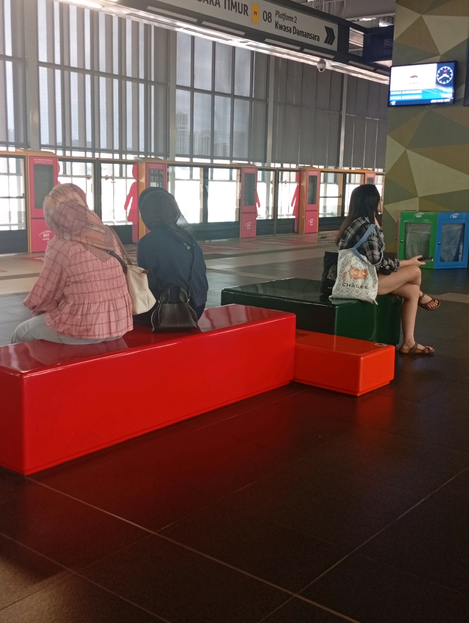The image depicts a platform at an MRT station in Petaling Jaya, Malaysia, with three individuals visible. In the foreground, two women are seated on a bright red bench. The woman on the left wears a pink plaid blouse and a hijab, and a light-colored purse rests beside her. To her right, another woman in a dark hijab sits with a dark handbag in front of her. Further back, a third woman with long dark hair, wearing a plaid shirt and shorts, sits on a dark green bench. She is holding a mobile phone and a tote bag with a graphic design is slung over her shoulder. The platform features red and orange benches, dark green seating, and a column with a geometric design. A digital display board above the platform indicates "Platform 2" and "Kwasa Damansara." The background shows the station's glass-paneled walls with vertical blinds and advertisements. Two recycling bins, one green and one blue, are positioned against the wall on the right side of the image.