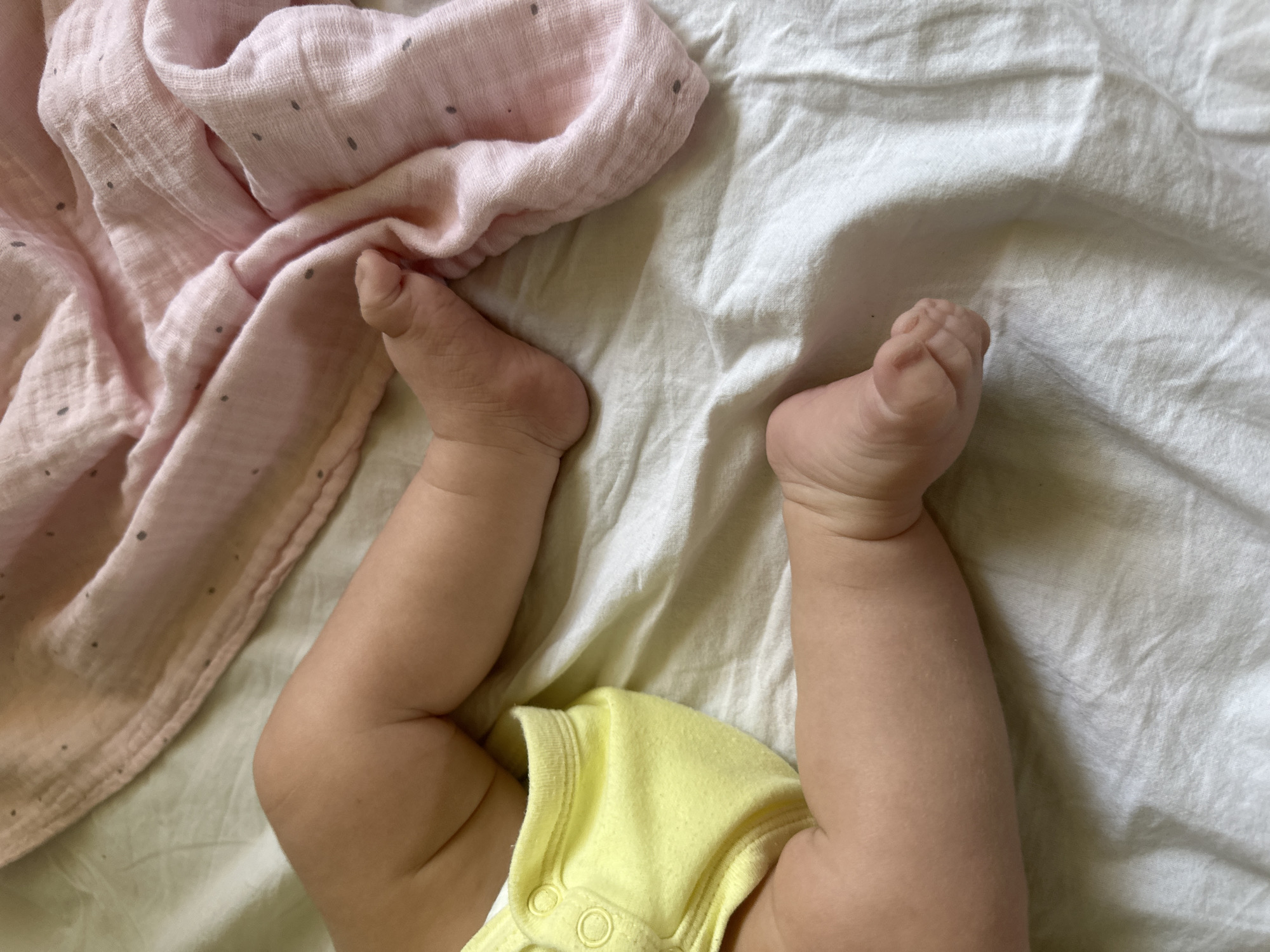 The image shows a close-up view of a baby's legs and feet. The baby is wearing a yellow onesie. To the left of the baby's legs, a pink muslin swaddle with small gray polka dots is draped over a white surface. The white surface appears to be a bedsheet or blanket, characterized by gentle folds and creases, suggesting a comfortable indoor setting. The lighting suggests it might be daytime, possibly indoors with natural light. The image conveys a sense of tenderness and domesticity, focusing on the soft skin and delicate features of a young child. There is no visible text in the image. The location context provided is Plovdiv, Bulgaria.