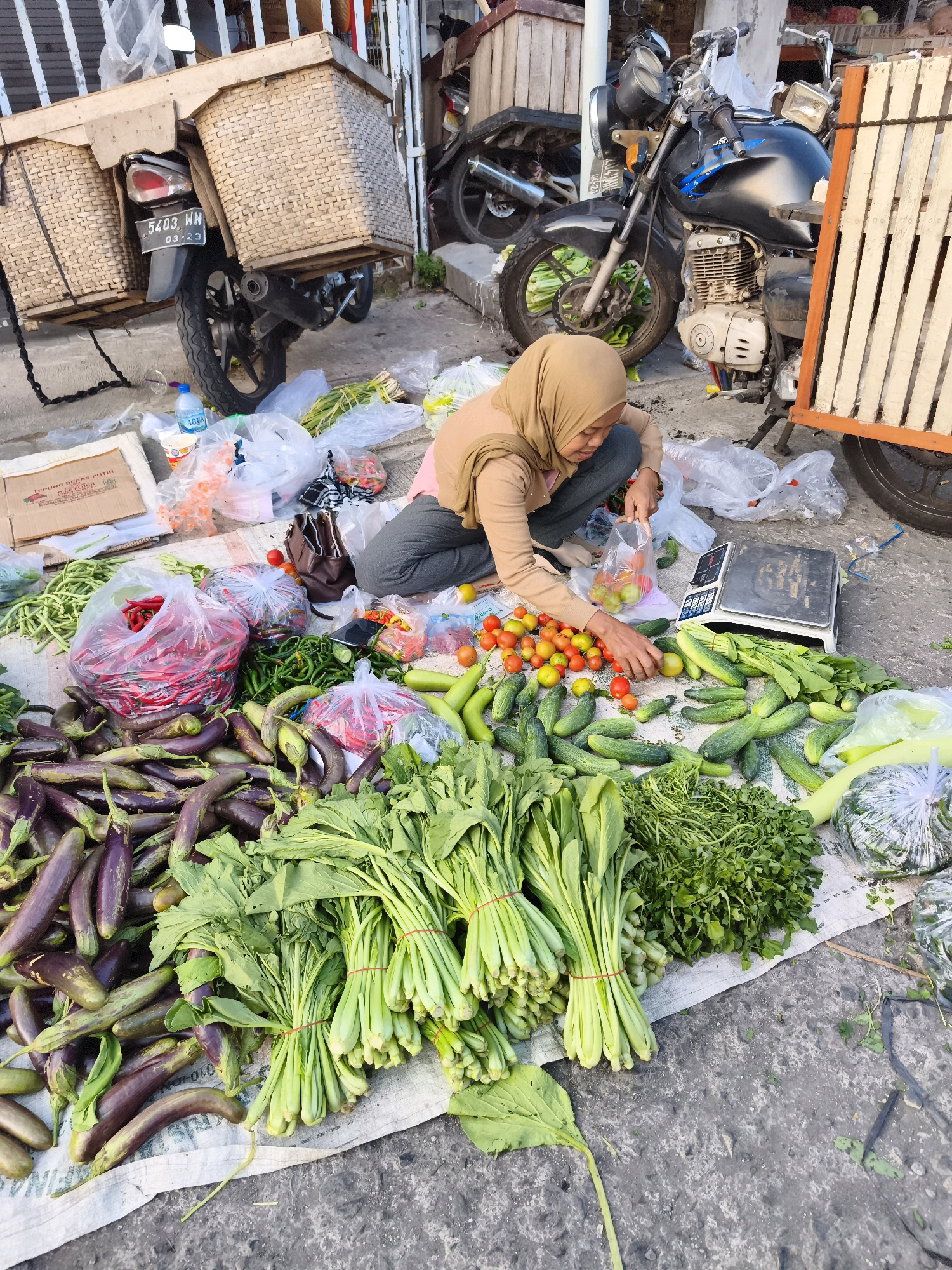 A female vendor wearing a light brown hijab and a beige top is kneeling on the ground in front of a display of fresh produce in Kaligondo, Indonesia. She is holding a plastic bag with red tomatoes and appears to be sorting or bagging them. The produce, including eggplants, cucumbers, green chilies, red chilies, tomatoes, and leafy greens, is spread out on a white cloth laid on the concrete ground. Behind her, a black motorcycle with large wicker baskets attached to its sides is parked. To the right, a wooden stall structure with vertical slats is visible, along with a digital scale placed on the cloth next to the produce. Several plastic bags, a bottle of "AQUA" water, and other assorted items are scattered around the vendor and the vegetables. The scene suggests an outdoor market or roadside stall.