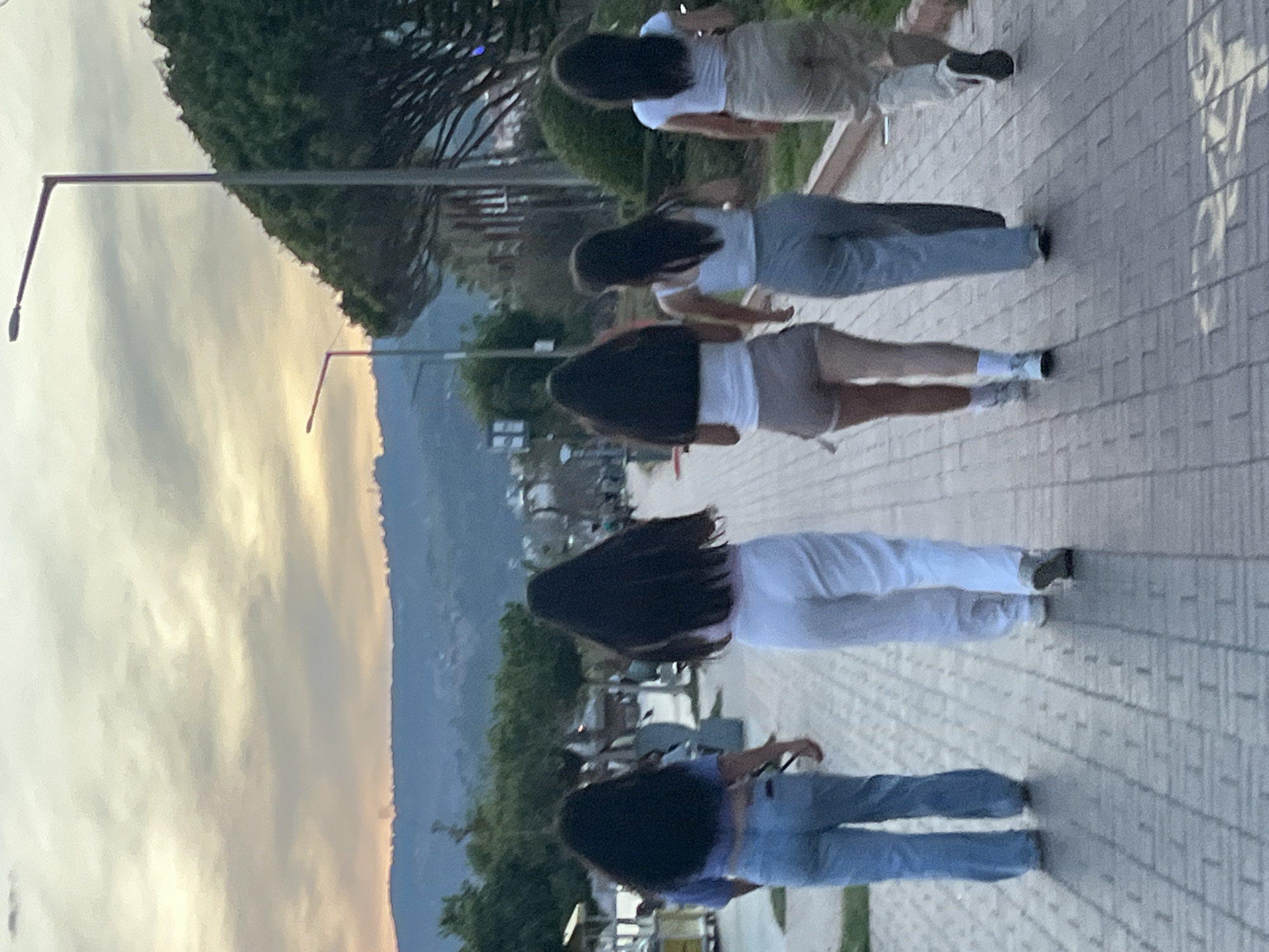 The image shows five people with long dark hair walking on a paved path in Alanya, Turkey. From right to left, they wear: a white top and tan shorts, a white top and light blue jeans, a white top and tan shorts, a white top and white pants, and a purple top and light blue jeans. In the background are trees, buildings, and a mountain range under a cloudy sky at sunset. A street light is visible on the left.