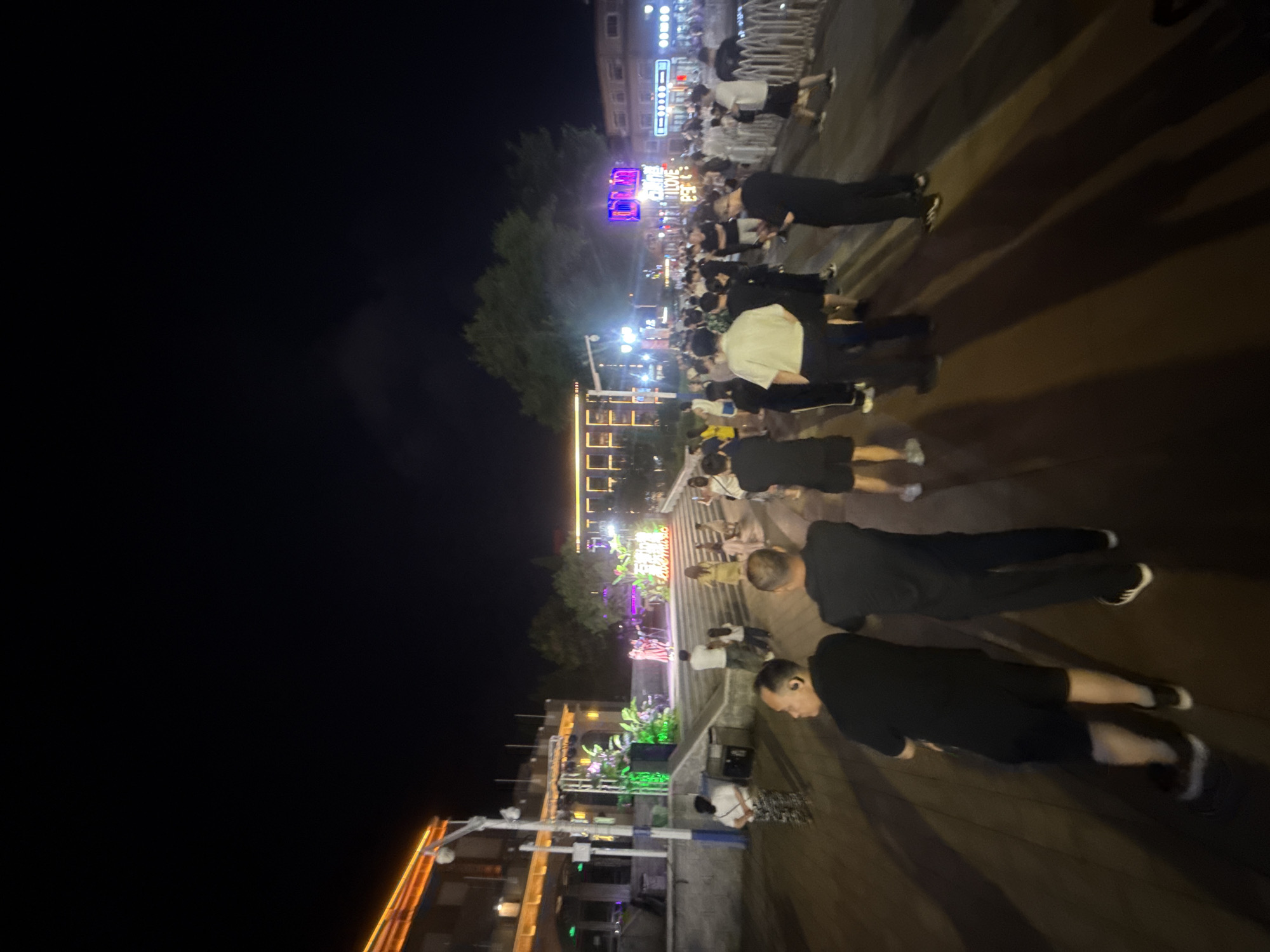 This image captures a lively night scene at a waterfront promenade in Qinhuangdao, China. A group of people are gathered, some facing a stage where a performance appears to be taking place. The stage is illuminated with green lights and decorated with plants, suggesting an outdoor event. Neon signs, including one that reads "LOVE MUSIC," are visible in the background, adding to the festive atmosphere. The ground is paved, and shadows stretch across it, indicating nighttime lighting. The people are dressed casually, and their presence suggests a public gathering or event. The architecture in the background appears to be modern, with multiple stories and illuminated windows.