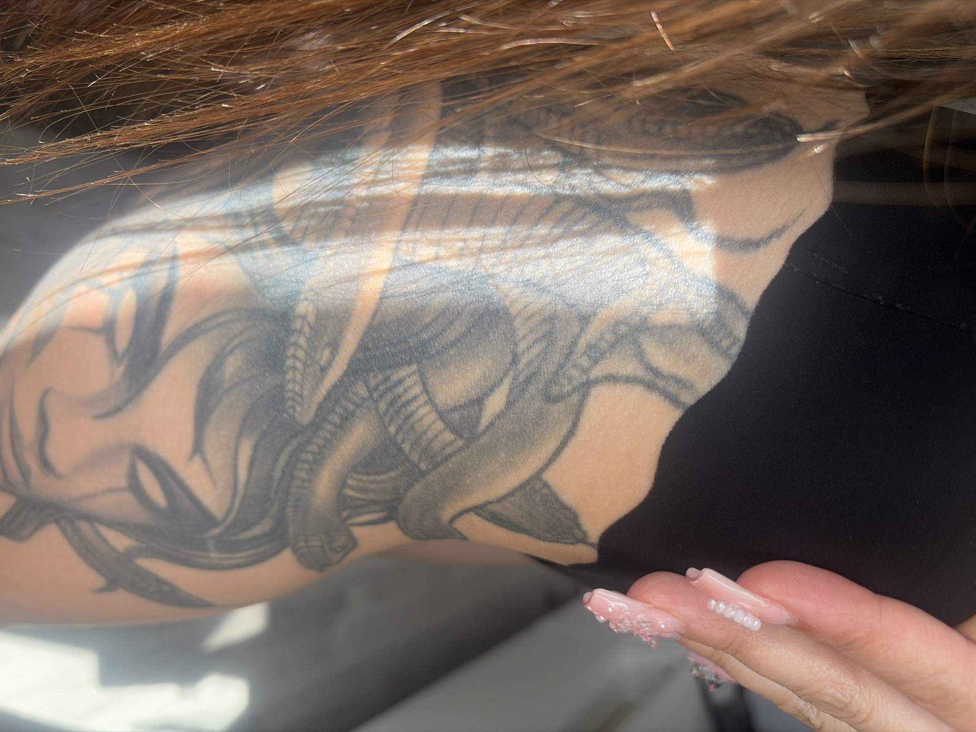 The image shows a close-up of a person's arm with a tattoo of Medusa. The tattoo is detailed, depicting Medusa's face with snakes for hair and a knife. The person's hair is long and reddish-brown, and their fingernails are decorated with pink gel and small white beads. The person is wearing dark clothing. Sunlight casts shadows on the arm, highlighting the tattoo. The background is blurred, but appears to be indoors with a neutral-colored wall and possibly furniture. The image does not provide any information about the location in the city of Saint Petersburg, Russia.