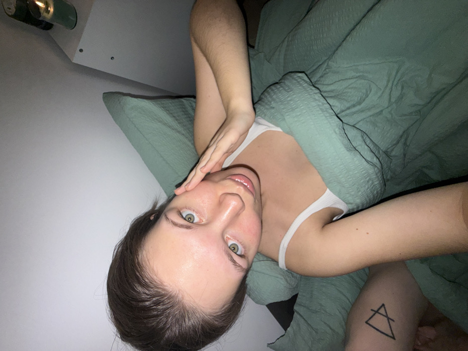 A woman with fair skin and green eyes, dressed in a white tank top, lies in bed under a green comforter. Her hair is dark brown and pulled back from her face. She has her hand near her face and is looking at the camera. A tattoo of a triangle with a horizontal line bisecting it is visible on her arm. A candle sits on a shelf in the background.