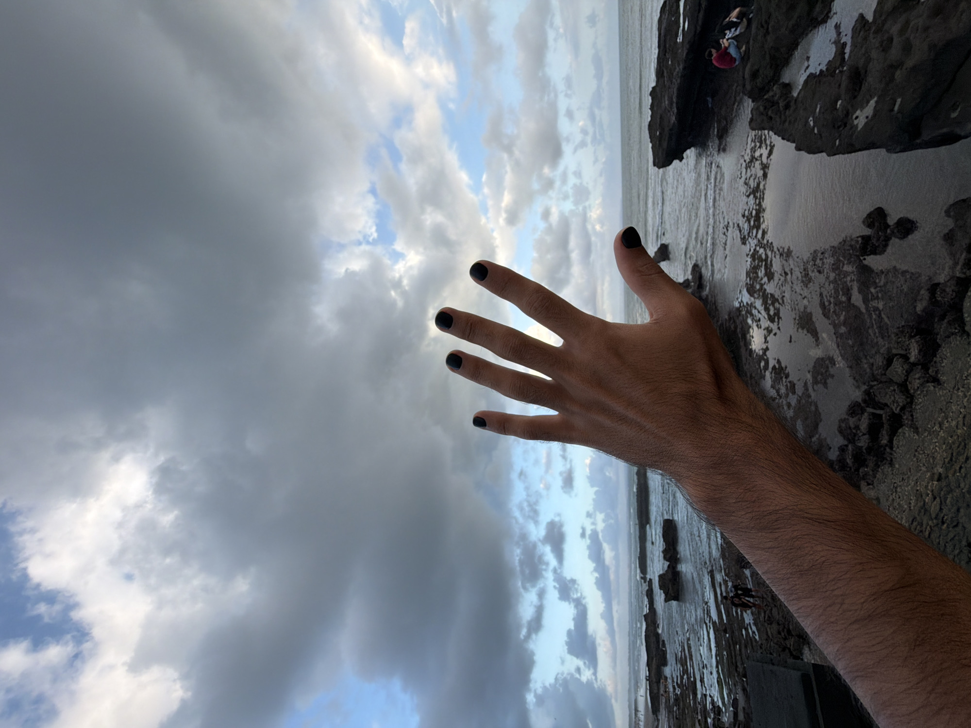 This media file captures a human arm and hand, extending into a cloudy sky, rotated clockwise. The hand, adorned with black nail polish, is the dominant subject in the foreground. The arm is visible up to the elbow, featuring fine hair.
The background reveals a coastal scene, likely a beach or rocky shoreline in North Kuta, Indonesia. To the right of the hand, the ocean meets a rugged, dark rocky shore. Small waves are visible in the water. Several other individuals can be seen on the rocks in the distance, appearing small and engaged in activities near the water's edge, possibly exploring or relaxing. One person in a red shirt is somewhat discernible perched on a rock.
The sky is filled with a mixture of dark, heavy grey clouds and brighter, more diffuse white clouds, with patches of clear blue sky peeking through. This suggests an overcast day, possibly nearing dusk given the soft, diffused light and the contrast between the dark clouds and the clearer sky. The overall atmosphere appears calm and natural. There is no visible text in the image.