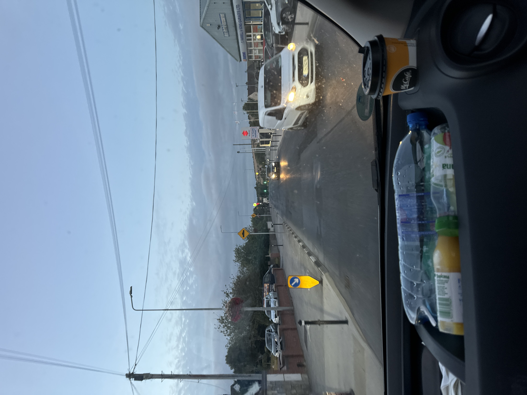 The image depicts a street view in Tramore, Ireland, likely captured from inside a vehicle. The scene is set during dusk, with a pale blue sky transitioning into darker hues, and scattered clouds. A street with a wet surface, suggesting recent rain, stretches into the distance. Various elements of an urban environment are visible: streetlights, power lines, and traffic signs, including a yellow diamond-shaped sign with a hump symbol and a blue directional arrow sign on the sidewalk.
Several cars are present: parked cars on the left, a dark car further down the road, and a white van with its headlights on in the right lane. The van appears to be a Ford Transit Custom. To the right of the road, a commercial building with a sign reading "T. FARRELL & SONS GARAGES LTD." is visible, along with other businesses, including a "SUZUKI" dealership displaying a "10 YEARS WARRANTY" sign.
In the foreground, inside the vehicle from which the photo is taken, a dark dashboard area is visible. On it rests a McCafe coffee cup and two plastic bottles, one filled with a clear liquid and the other with orange juice. The overall impression is that of a typical evening drive through a suburban commercial area in Tramore.