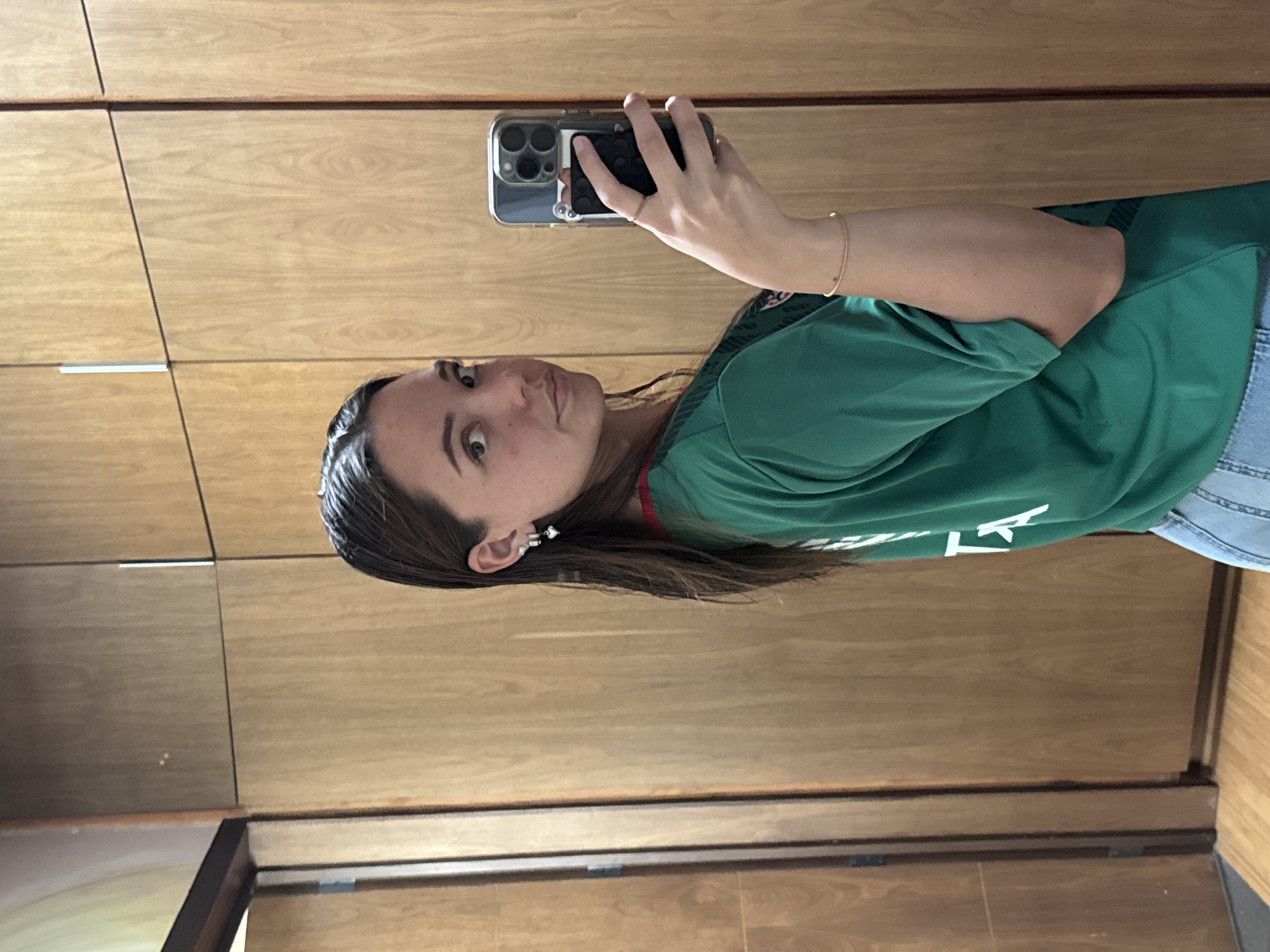 A woman is taking a selfie in front of a wooden wardrobe or wall paneling. She is wearing a green shirt with white lettering and what appears to be a red trim. She is holding a smartphone in her right hand. A gold bracelet is visible on her right wrist. The background suggests an interior space. The specific location of San Bernardino Tlaxcalancingo, Mexico cannot be determined from the visual information.