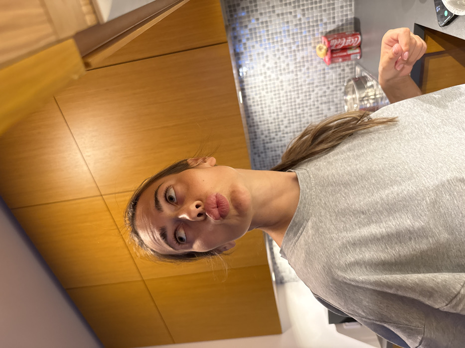 This image captures a female subject from the mid-torso up, situated in a modern kitchen. The subject, with light brown hair pulled into a ponytail, wears a loose grey t-shirt. She exhibits a playful expression, with her head tilted slightly to the right, eyes gazing upward and to the left, and lips pursed into a "kissing" or "fish face" pose. Her right hand, partially clenched with light pink nail polish, is visible at the lower right of the frame.
The background features light brown wooden upper cabinets spanning the width of the scene, with under-cabinet lighting illuminating a backsplash of small, light-colored square tiles. A dark countertop is visible beneath the backsplash. On the countertop to the right of the subject, two red Coca-Cola cans are stacked, topped by a light-colored ring-shaped object. A metallic bowl or pot is partially visible to the subject's left, while a sliver of a white dish and a dark smartphone are present on the far right of the countertop. The environment appears to be a well-lit residential kitchen. The image itself provides no identifiable landmarks or specific details to determine a precise location within the city of Warsaw, Poland.