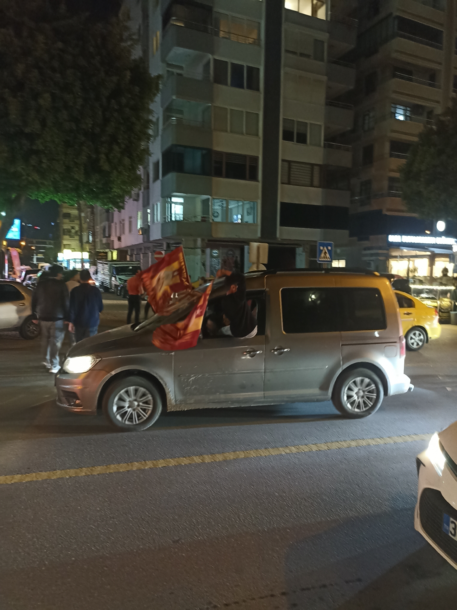 Scene in Yenişehir, Turkey at night: A silver van with a flag (red and gold) emerging from the driver's side window has one or more occupants. The street is paved and lined with yellow paint, buildings (grey and white). Pedestrians (including some wearing dark jackets) stand on the street near the van. Also visible: a yellow car, and the edge of a white car.