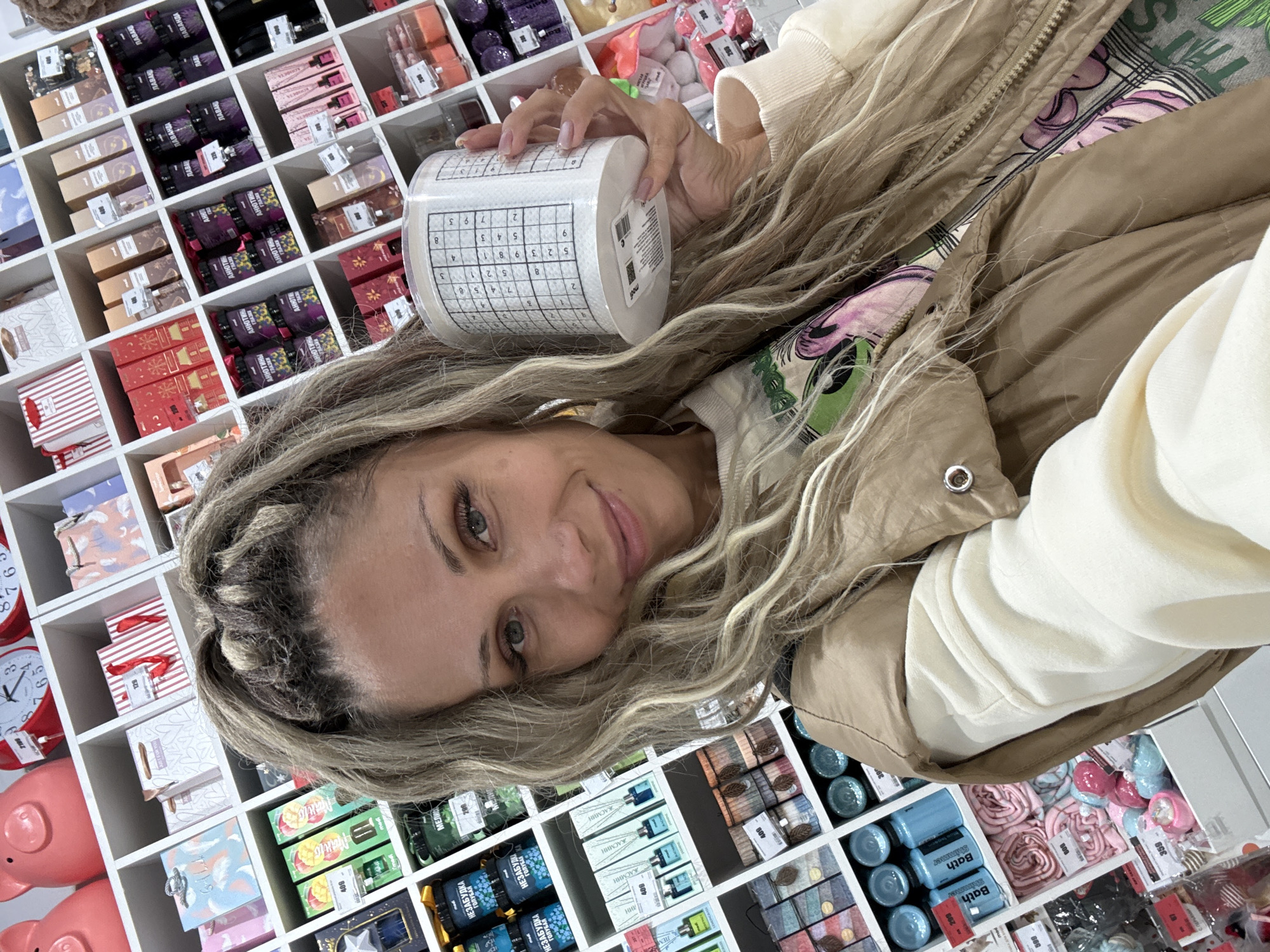 A blonde woman with braided hair is taking a selfie in a store. She is smiling and holding a roll of toilet paper with a grid pattern on it. She is wearing a beige vest over a cream-colored sweatshirt with a graphic print. The background is a wall of shelves filled with various products, including gift bags, jars, bottles, and decorative items. The lighting is bright and appears to be indoor store lighting. The location is not discernible from the image.