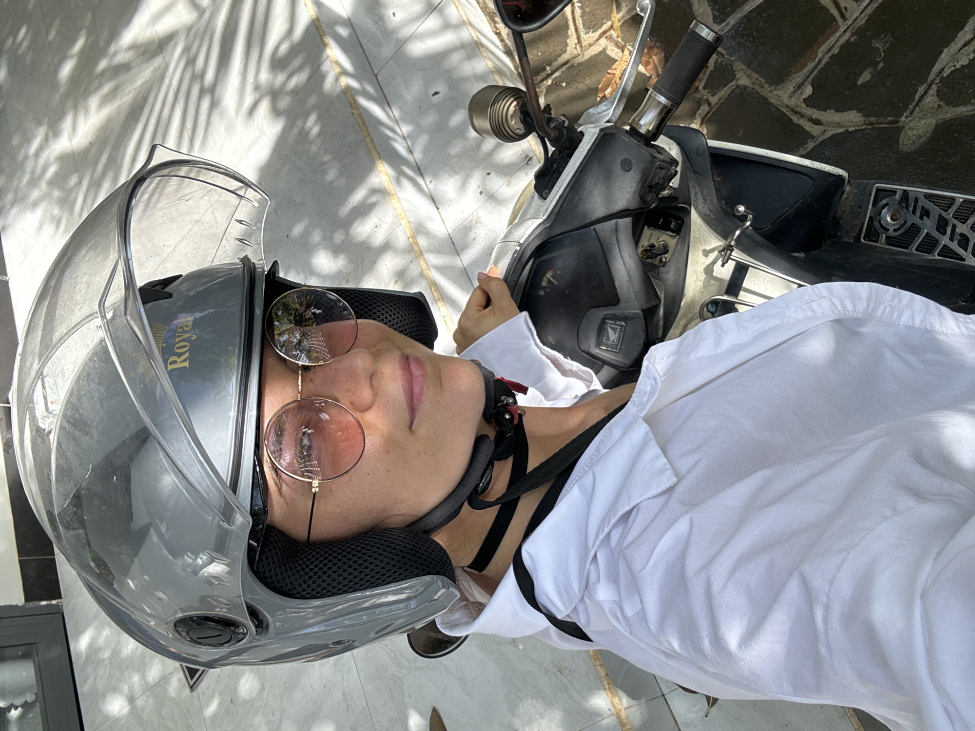 A person wearing a silver helmet with a clear visor and round-framed sunglasses is positioned on or next to a scooter. The individual is also wearing a white shirt. The scooter is parked on a paved surface with shadows cast by vegetation. The location is likely Mũi Né, Vietnam.