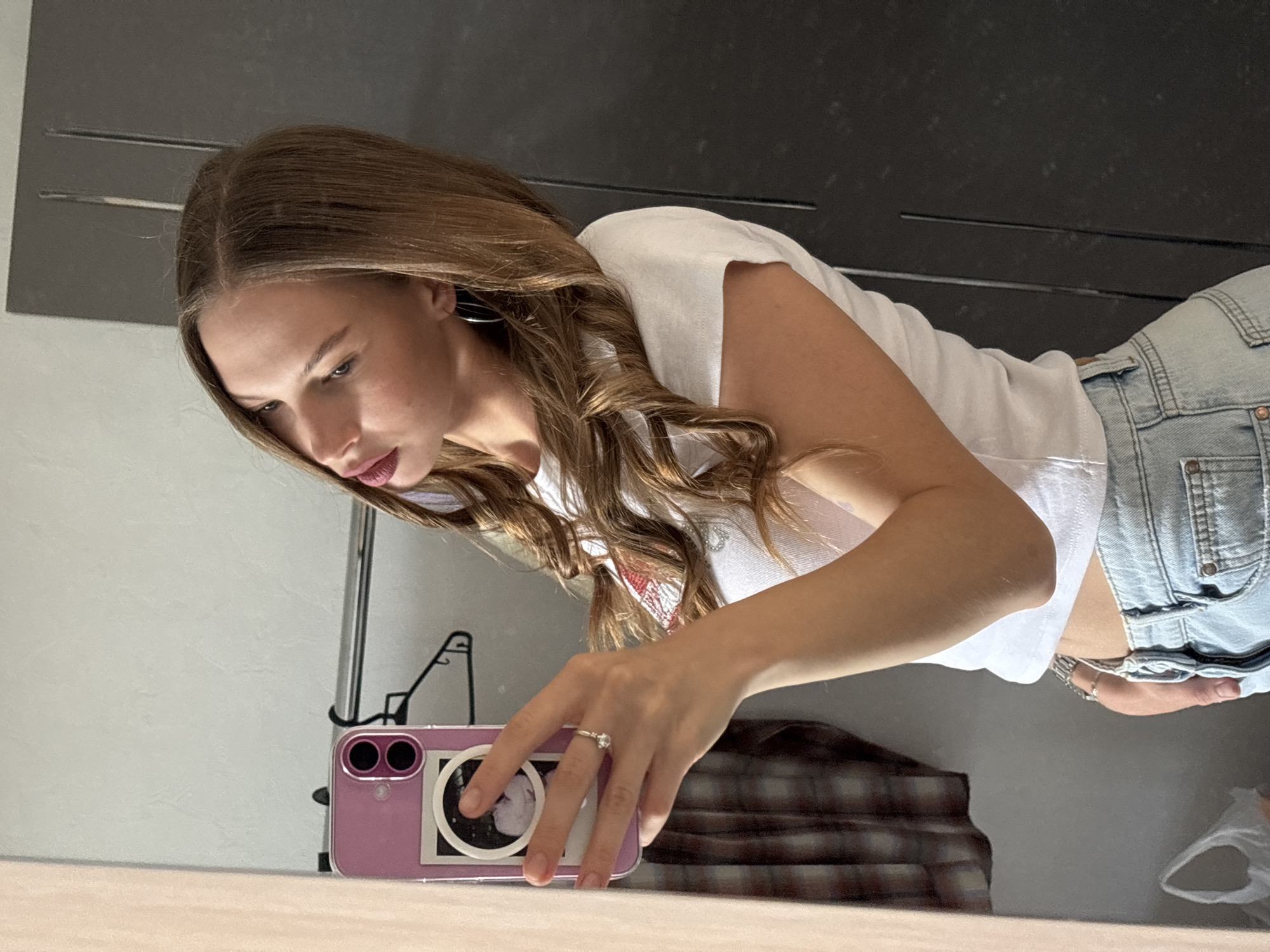 A young woman with long, wavy brown hair is captured in a mirror selfie. She is wearing a white t-shirt and light-wash denim shorts. Her gaze is directed downwards towards a pink smartphone she is holding in her right hand, which is resting on a table. A silver ring with a prominent stone adorns her ring finger. The background suggests an indoor setting, possibly a bathroom or dressing area, with a large reflective surface (likely a mirror) dominating the upper portion of the image. Hints of a light-colored wall and a dark plaid fabric are visible. The overall mood appears casual and personal. There is no visible text in the image.