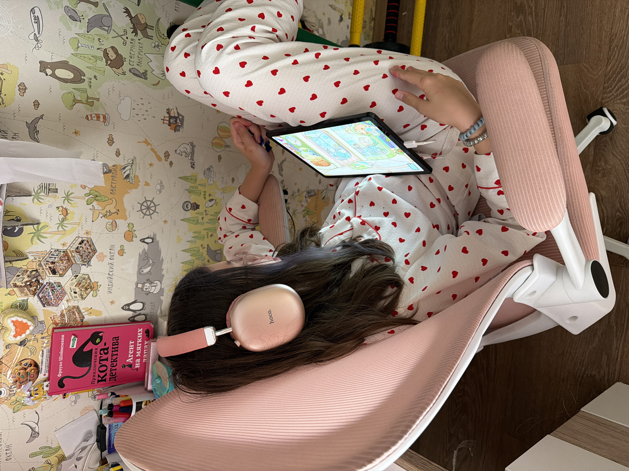 A young person is lying on their side on a pink chair, looking at a tablet computer. They are wearing white pajamas with red hearts and pink headphones. The tablet is displaying a colorful cartoon. Next to the person, on a colorful mat with a world map design, is a pink book with a black cat on the cover. The book has Russian text that translates to "Adventures of a Cat Detective: Agent on Soft Paws". There are also some pens, a cookie, and a heart-shaped object on the mat. The scene appears to be indoors, possibly in a bedroom or play area. The lighting is bright, suggesting it could be daytime. The presence of a children's mat and pajamas suggests the subject is a child or adolescent.