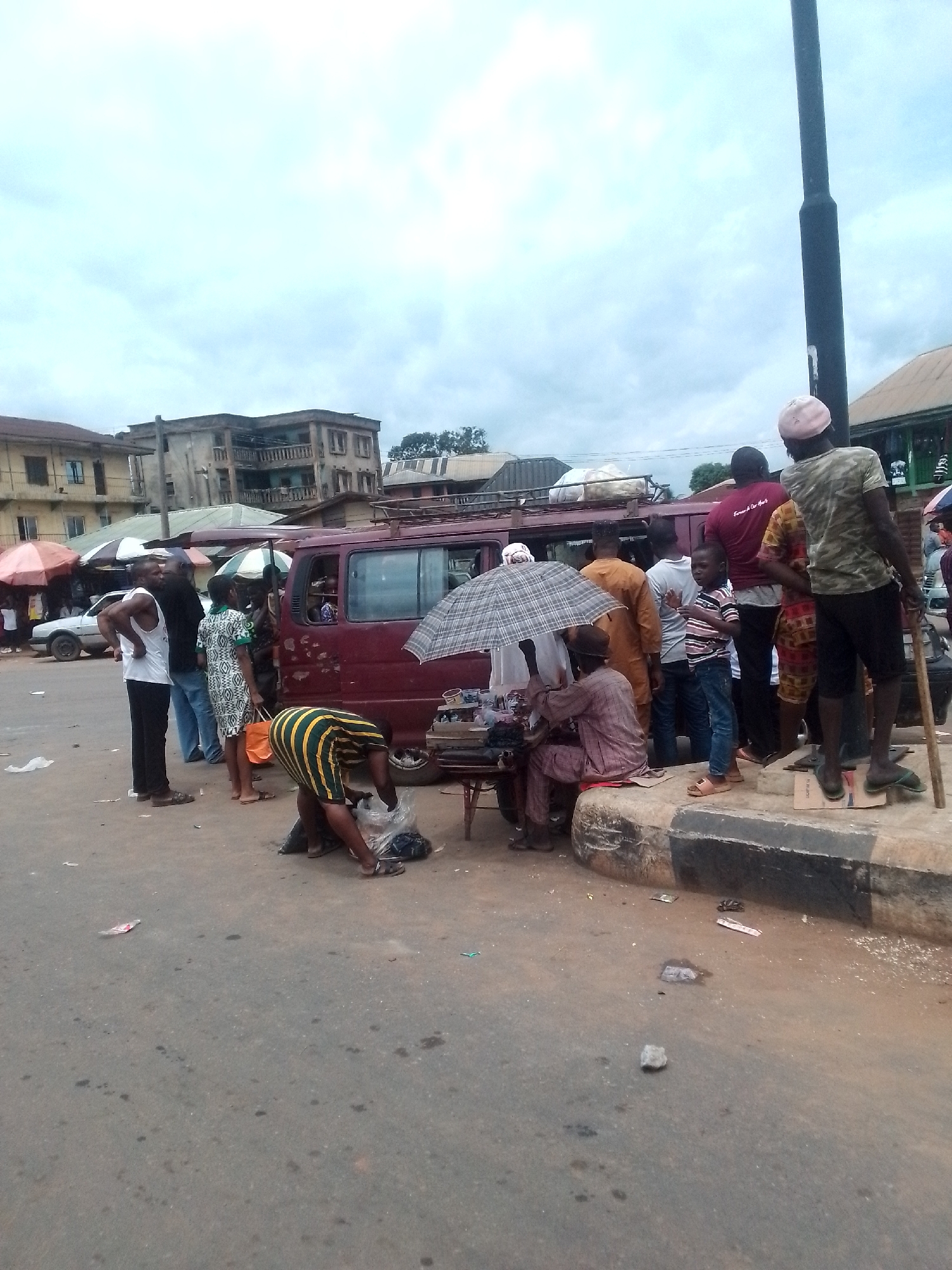 The image shows a street scene in an urban location in the city of Unknown, Unknown. A maroon van with luggage on its roof is parked on the side of the road, and several people are standing near it. Some appear to be boarding the van, while others are waiting. There is a vendor seated with an umbrella overhead, possibly selling goods from a small table. A person is bending down, appearing to interact with something on the ground. Litter and debris are scattered on the road surface. Buildings are visible in the background, and a utility pole stands on the sidewalk. The sky is overcast.