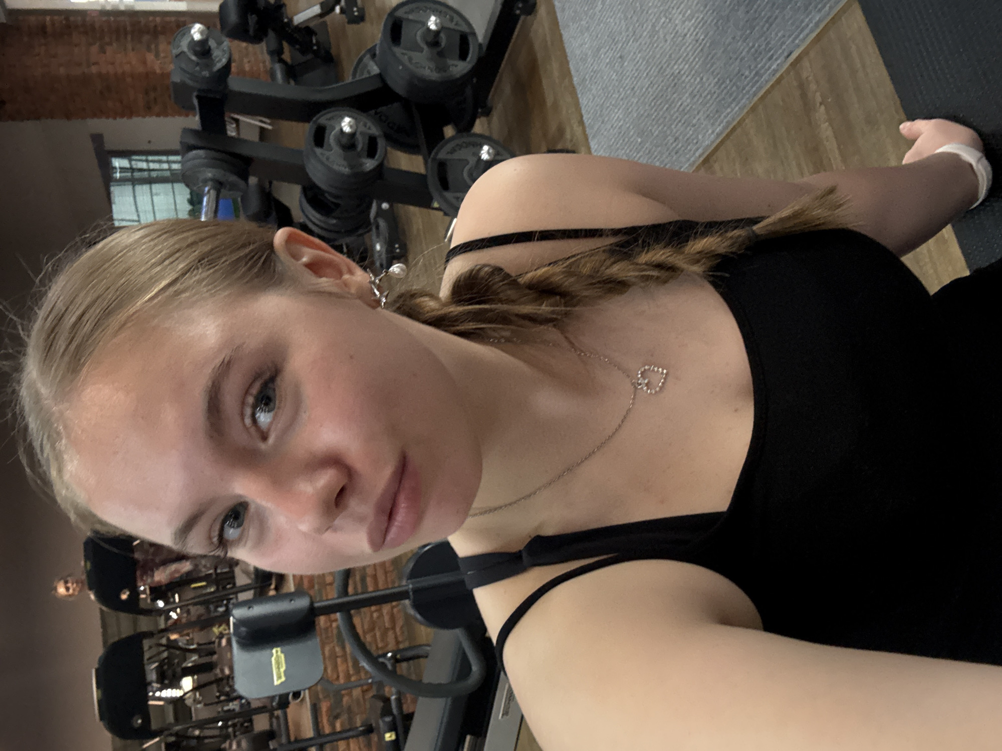 The image depicts a young woman in a gym, likely in the city of Chaikovsky, Russia. She has light blonde hair pulled back in a braid, and she is wearing a black tank top with thin straps and a silver heart-shaped pendant necklace. She is fair-skinned. In the background, gym equipment, including weight racks and exercise machines, is visible. A portion of a gray mat is also in the frame. The perspective suggests she is holding the camera or phone herself, as it is a selfie.