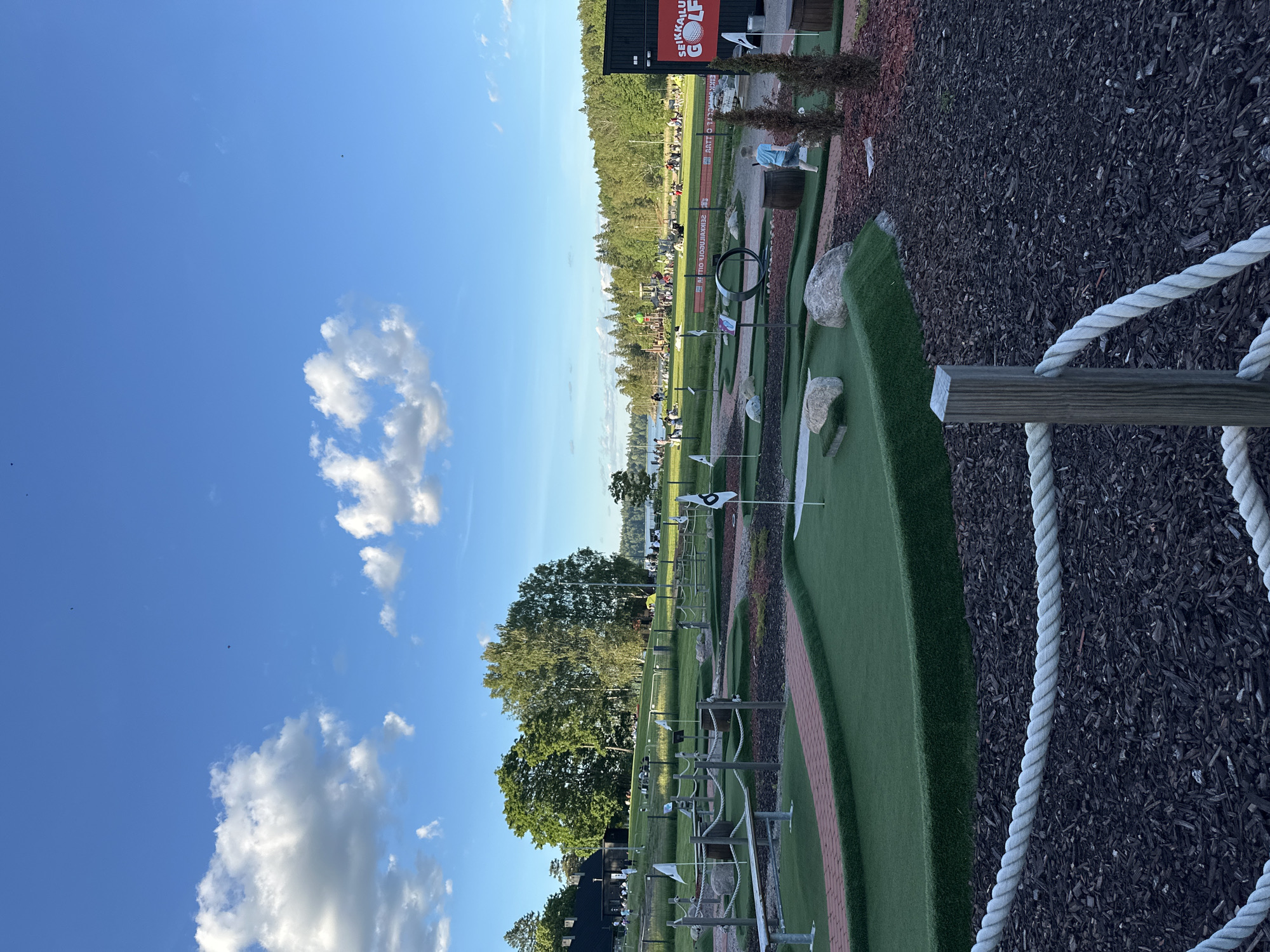 The image depicts an outdoor mini-golf course on a sunny day. The course features artificial turf, brick pathways, and various obstacles, including barrels, rocks, and a metal hoop. Several flags are visible throughout the course, marking the holes. In the background, there are trees, a building, a body of water, and a playground area with people scattered around. The sky is blue with scattered white clouds. A young boy in a blue shirt is walking on the course. A sign that reads "SEIKKAILU GOLF" is visible on a dark building to the right. The foreground is dominated by a wooden post with a thick white rope acting as a barrier, set against a backdrop of dark wood chips.