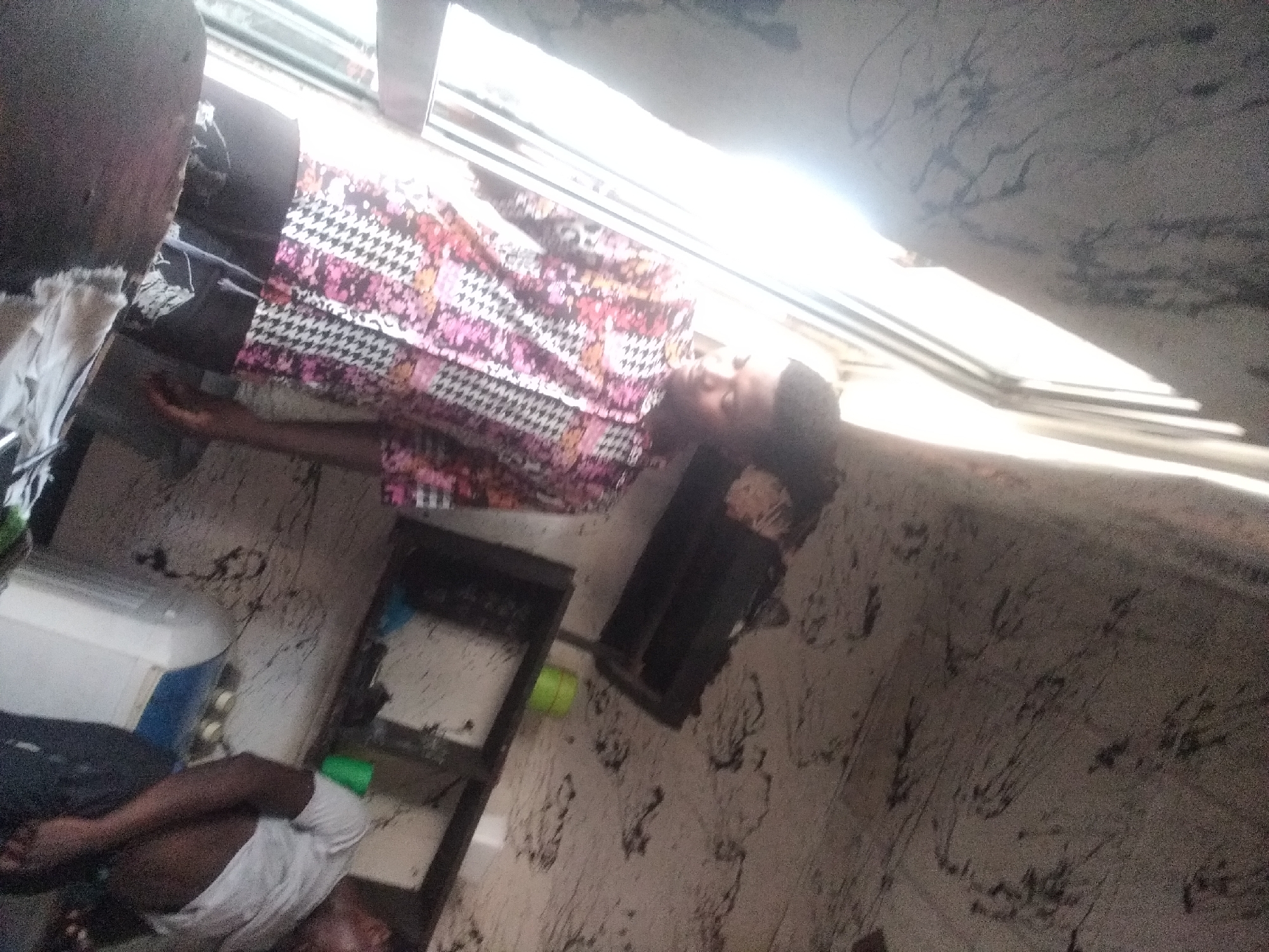 The image depicts an indoor scene with two individuals. The foreground features a young man wearing a patterned shirt and dark trousers, partially obscured by a dark object. His posture suggests he is looking downwards. Behind him, a second individual, wearing a white t-shirt, is visible, also with their head down. The environment appears to be a room with a large window or door letting in bright light, creating a stark contrast between light and shadow. Shelving units are present on the wall, holding various items, including a green container. The walls and ceiling show evidence of discolored marks. The scene is set in Jalingo, Nigeria.