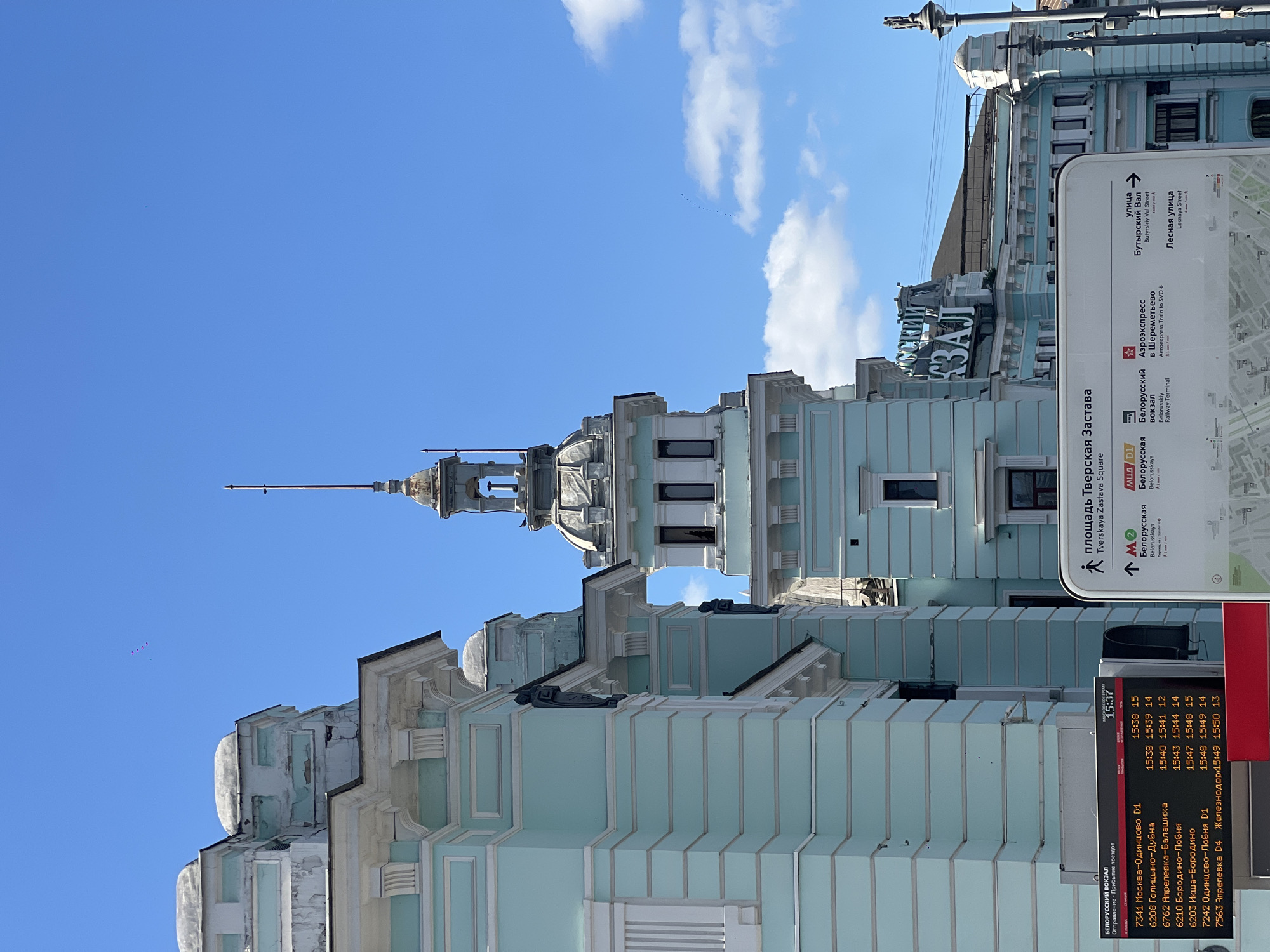 The image shows a portion of a light blue building with ornate architectural details and a spire. Attached to the building is a signpost, partly in Russian and partly in English. The signpost includes a map, directions, and transportation information, specifically about Belorussky Railway Terminal and Aeroexpress to Sheremetyevo Airport. The timetable displays departures and arrivals for trains, including destinations such as Moscow, Odintsovo, Dubna, Balashikha, and Lobnya. The signpost identifies the location as "площадь Тверская Застава" or Tverskaya Zastava Square in Moscow, Russia. The sky is blue with some white clouds.