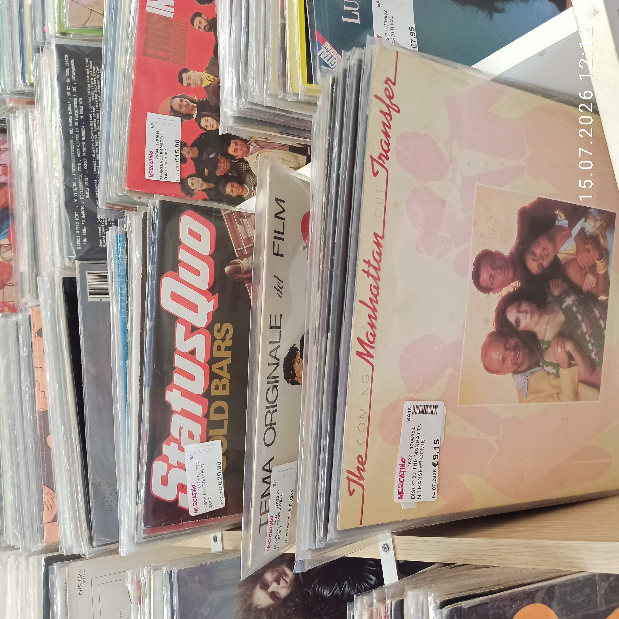 The image shows a close-up of a retail display of vinyl records. The records are mostly in protective plastic sleeves, with some of their album art visible. Several labels with "MERCATINO" are affixed to the sleeves, indicating a second-hand market or a specific type of store.
One prominent album visible is by "Status Quo" titled "GOLD BARS". Another is labeled "TEMA ORIGINALE del FILM" and features a portrait of a man. Further down, an album by "The Coming Manhattan Out Transfer" is displayed with a photograph of four people, two men and two women, posed together. There are also partial views of other albums, including one with "EROS RAMAZZOTTI" visible on its cover.
Prices and dates are visible on some of the Mercatino labels: €20,00 with a date of 26.03.2026 for the Status Quo album, €17,00 with a date of 17.06.2026 for the film theme album, and €9,15 with a date of 04.07.2026 for the Manhattan Out Transfer album. Another label shows €7,95.
The overall environment appears to be an indoor retail space, possibly a record store or flea market, with the records organized on shelves or racks. The lighting is consistent with artificial indoor lighting. There is no specific indication of the city of Molfetta, Italy, in the visible elements of the image.