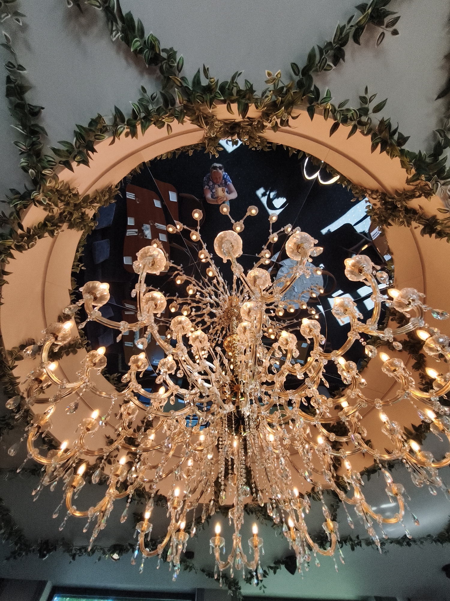 The image shows a grand, ornate crystal chandelier hanging from a ceiling with an arched opening. The opening is decorated with artificial greenery. Within the arched opening, a reflective surface acts as a mirror, showing a ceiling with decorative lights and a person wearing a blue and white patterned shirt, holding a small object. The chandelier is made of gold-colored metal and is adorned with numerous crystal droplets and flower-shaped light fixtures. The overall impression is one of opulent decor. This scene is located at the "Kalamaja Rõõm" cafe in Tallinn, Estonia.