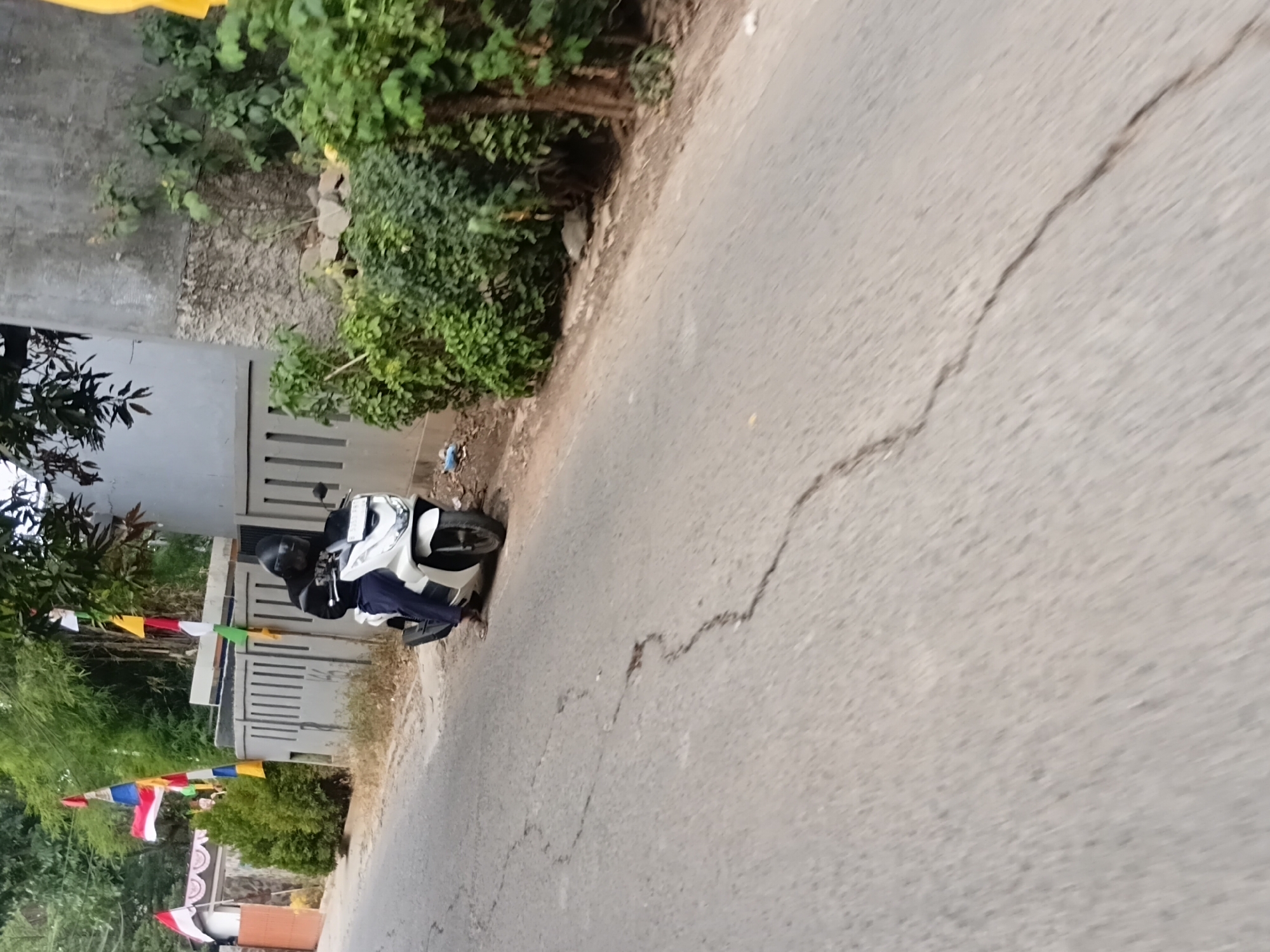 A person wearing a black helmet and black jacket is sitting on a white scooter on the side of a paved road in Setu, Indonesia. The scooter is parked next to a grey concrete fence and lush green foliage. Indonesian flags are visible hanging from poles along the fence line. The road surface shows signs of wear with several cracks. The background features a concrete building and trees.