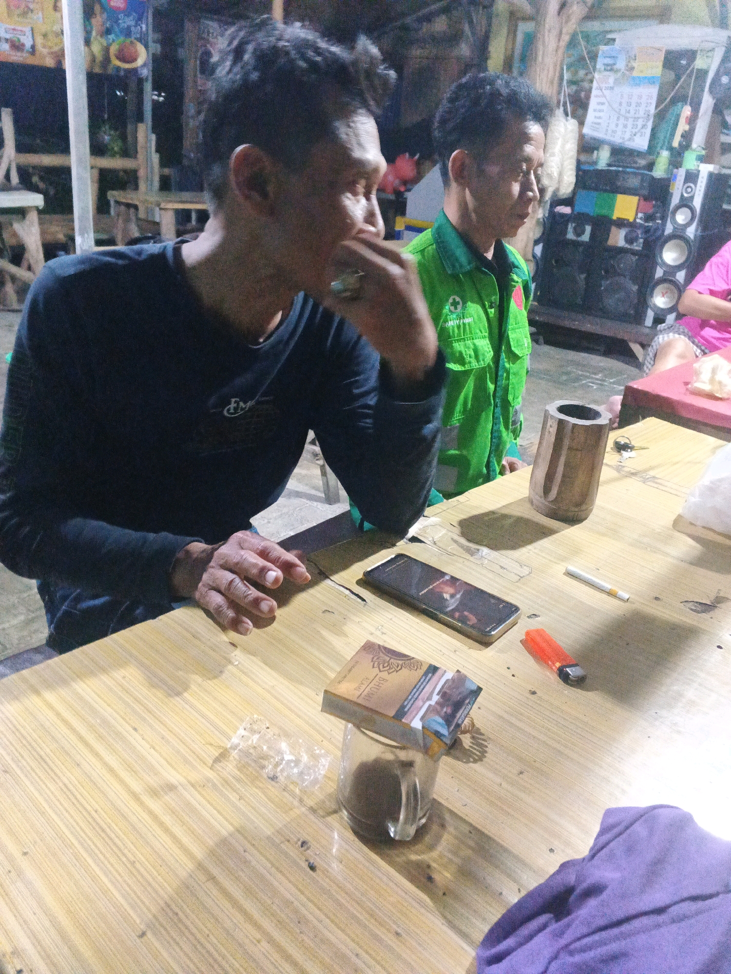 The image depicts an outdoor scene at night, possibly a roadside stall or eatery in Ungaran, Indonesia. Two men are present. The man in the foreground, wearing a dark long-sleeved shirt, is looking to his left, with his right hand near his mouth. The man in the background, wearing a bright green jacket over a dark shirt, is looking forward. On a wooden table in front of them are several items: a smartphone, a cigarette, an orange lighter, a pack of cigarettes, a glass with a dark liquid (likely coffee or tea), and a wooden cylindrical object. The table surface is illuminated by overhead lighting, casting shadows. In the background, there are indistinct structures, possibly buildings or stalls, and some electrical equipment like speakers. A calendar is visible on a wall in the background.