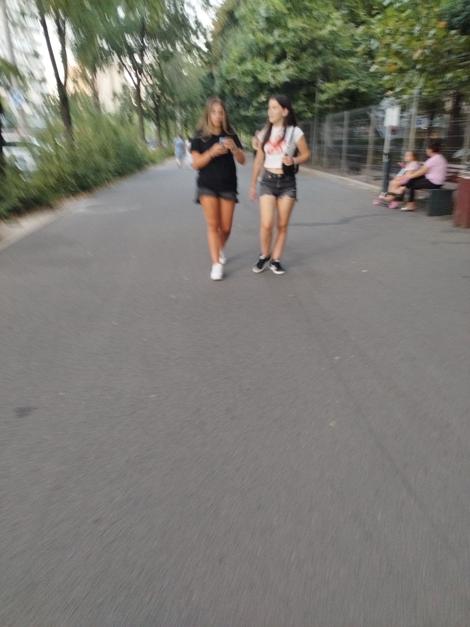 The image depicts an outdoor scene in Bucharest, Romania, featuring a paved pathway bordered by greenery and trees. Two young women are walking towards the viewer on the pathway. The woman on the left, with long brown hair, wears a black t-shirt and black shorts, and is holding a smartphone in her right hand. The woman on the right, with dark hair, is wearing a white t-shirt with a graphic print and denim shorts, and is carrying a black shoulder bag. In the background, other individuals are visible, including a person sitting on a bench and two people on a pink wheeled device, possibly a stroller or a cart. The environment suggests a park or a public walkway within the city. The overall image has a motion blur effect.
