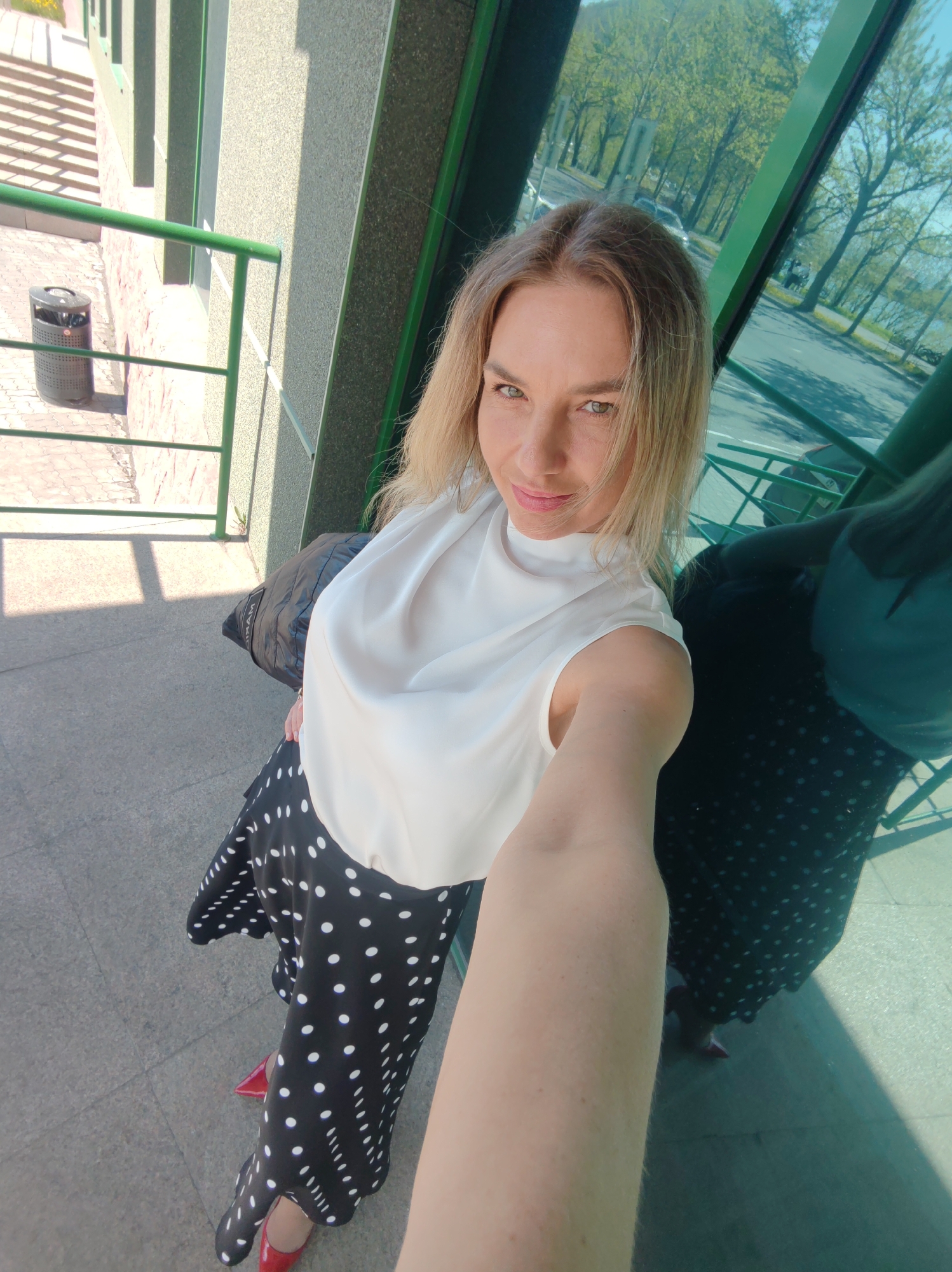 A blonde woman wearing a white sleeveless top and a black and white polka dot skirt takes a selfie outdoors. She is standing on a paved walkway next to a building with a green railing and large reflective windows. The woman's right arm and hand, holding the camera, are prominently featured in the foreground. Her reflection is visible in the window. In the background, trees and a street with cars can be seen. To the left of the woman, a trash can and a set of outdoor stairs are visible. The overall lighting suggests a sunny day.