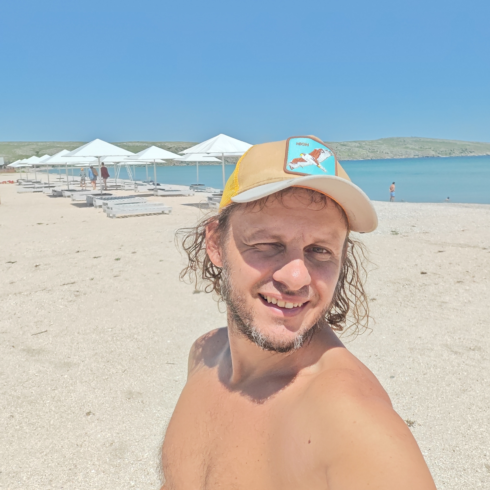 A man is standing on a beach in Мысовское сельское поселение, Ukraine. He is wearing a tan baseball cap with a bird emblem and is shirtless. The beach has white sand, white umbrellas, and lounge chairs. In the background, there is the sea, a coastline, and a clear blue sky. Two people are standing in the water, one further out than the other.