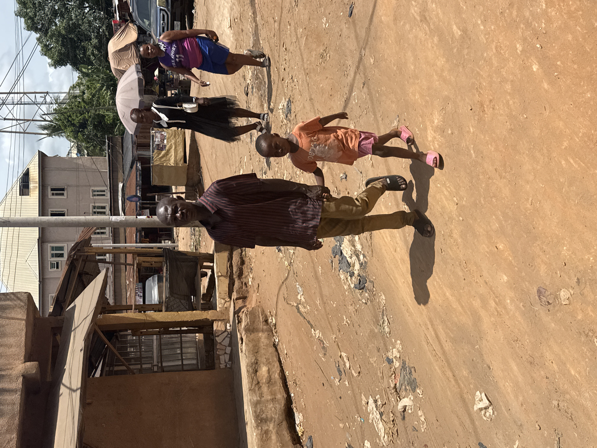 In Nkpor, Nigeria, the image captures an outdoor scene on a dirt road. A man in a striped shirt and khaki pants walks alongside a child in an orange shirt and pink shorts. Further up the road, a woman holds an umbrella and another woman wears a purple and blue dress. Buildings and power lines are visible in the background. Trash litters the side of the road.