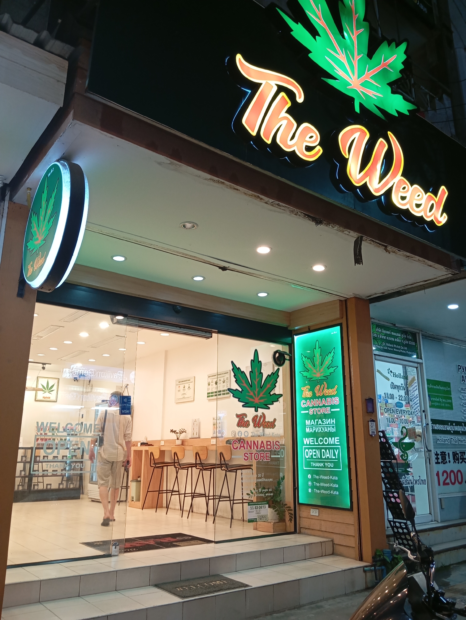 The image shows the exterior of a cannabis store named "The Weed" in Karon, Thailand. The store has a large, illuminated sign with a cannabis leaf and the store's name in neon lights. The storefront features a glass entrance and windows, with a welcome mat visible on the steps. Inside, a man wearing a light-colored shirt and shorts is walking away from the camera. The interior appears to be well-lit, with displays and seating visible. The street in front of the store is dimly lit, and a motorcycle is parked to the right of the frame. Signs in both English and Thai are present, indicating the store's location and operating hours.