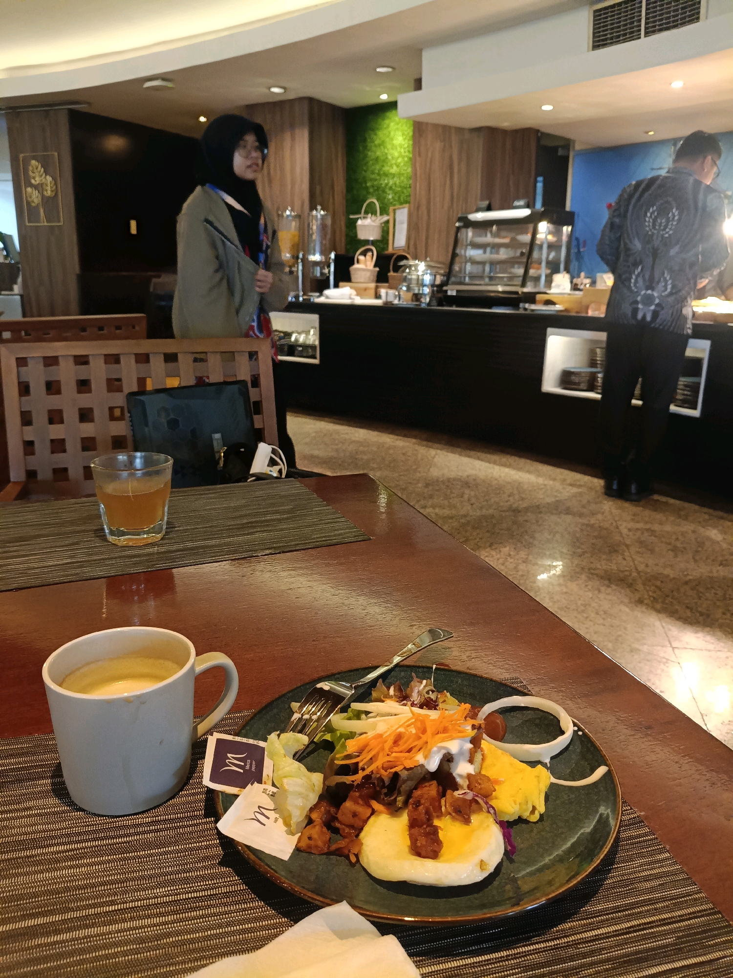 A dimly lit indoor scene, likely a hotel breakfast buffet in North Jakarta, Indonesia, shows a woman in a hijab and a man in a patterned shirt. In the foreground, a dark wood table holds a grey coffee mug filled with a creamy beverage, a small glass of amber liquid, and a plate of food. The plate contains a salad with shredded carrots, lettuce, and what appears to be diced meat and scrambled eggs, garnished with a swirl of white sauce and a slice of onion. Two small packets, one labeled "M" with "Moka coffee" and another with "Salt Pepper," rest near the plate. Behind the table, a patterned chair is visible. The background reveals a buffet counter with food displays, juice dispensers, and a green moss-like decorative wall. The floor is tiled with a reflective surface.