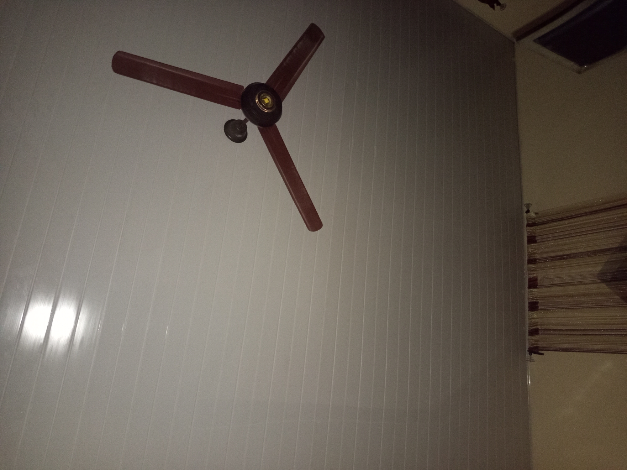 A ceiling fan is mounted on a white, paneled ceiling in a room. The fan has three brown blades and a central dark brown housing with gold accents. A bright light source is reflected on the ceiling to the upper left of the fan. Below the ceiling, a curtain with vertical stripes is visible, partially obscuring the wall. The scene appears to be an indoor residential space in Jalingo, Nigeria, based on the context provided.
