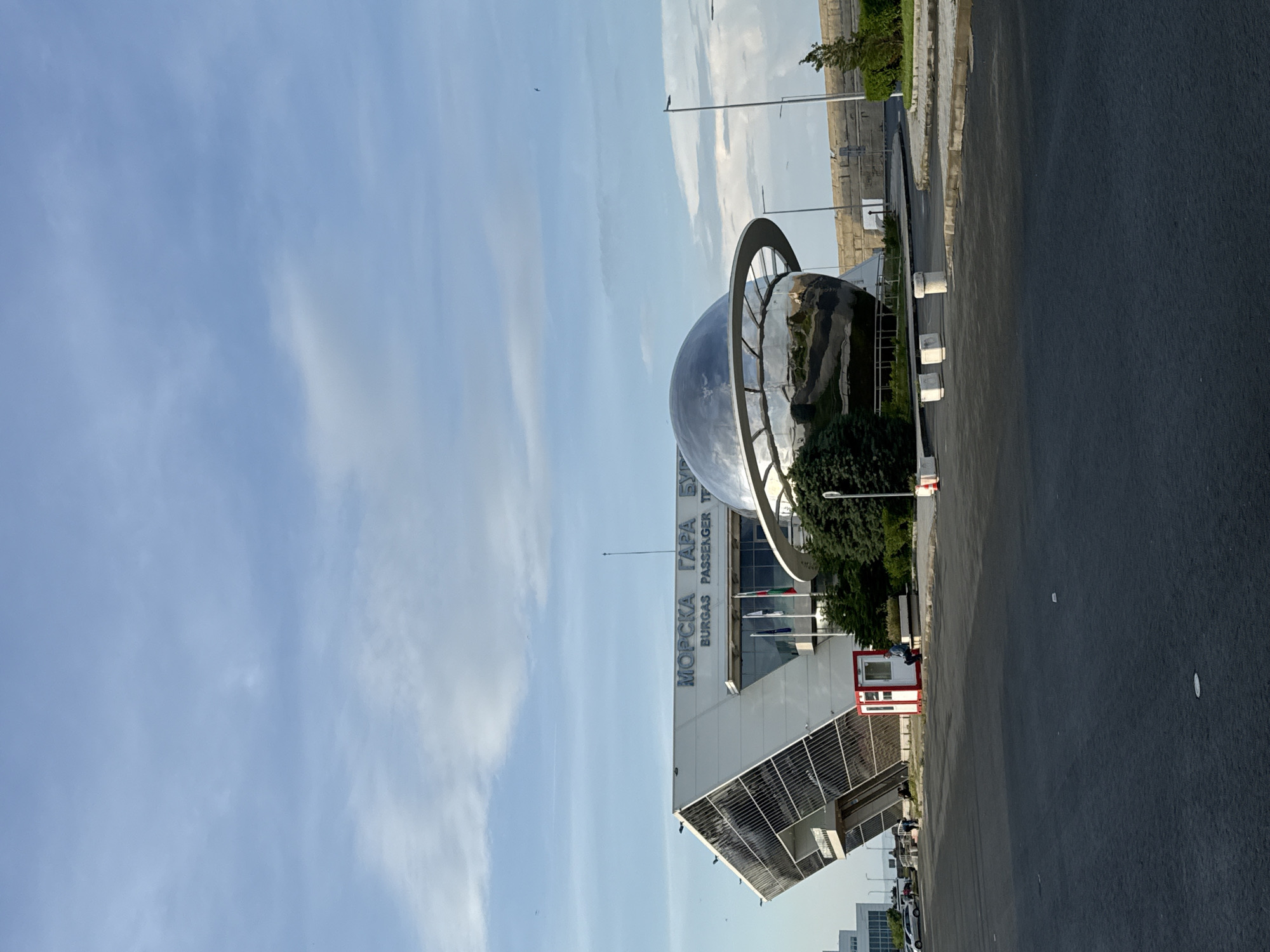 The image shows the Burgas Passenger Terminal in Burgas, Bulgaria. The building is modern and white, with large windows. A spherical metallic sculpture stands in front of the terminal. Several flagpoles display Bulgarian and other flags. A red and white booth is visible to the side. A paved area stretches in front of the terminal, and the sky is blue with sparse clouds. There are few people visible. A streetlamp is visible.