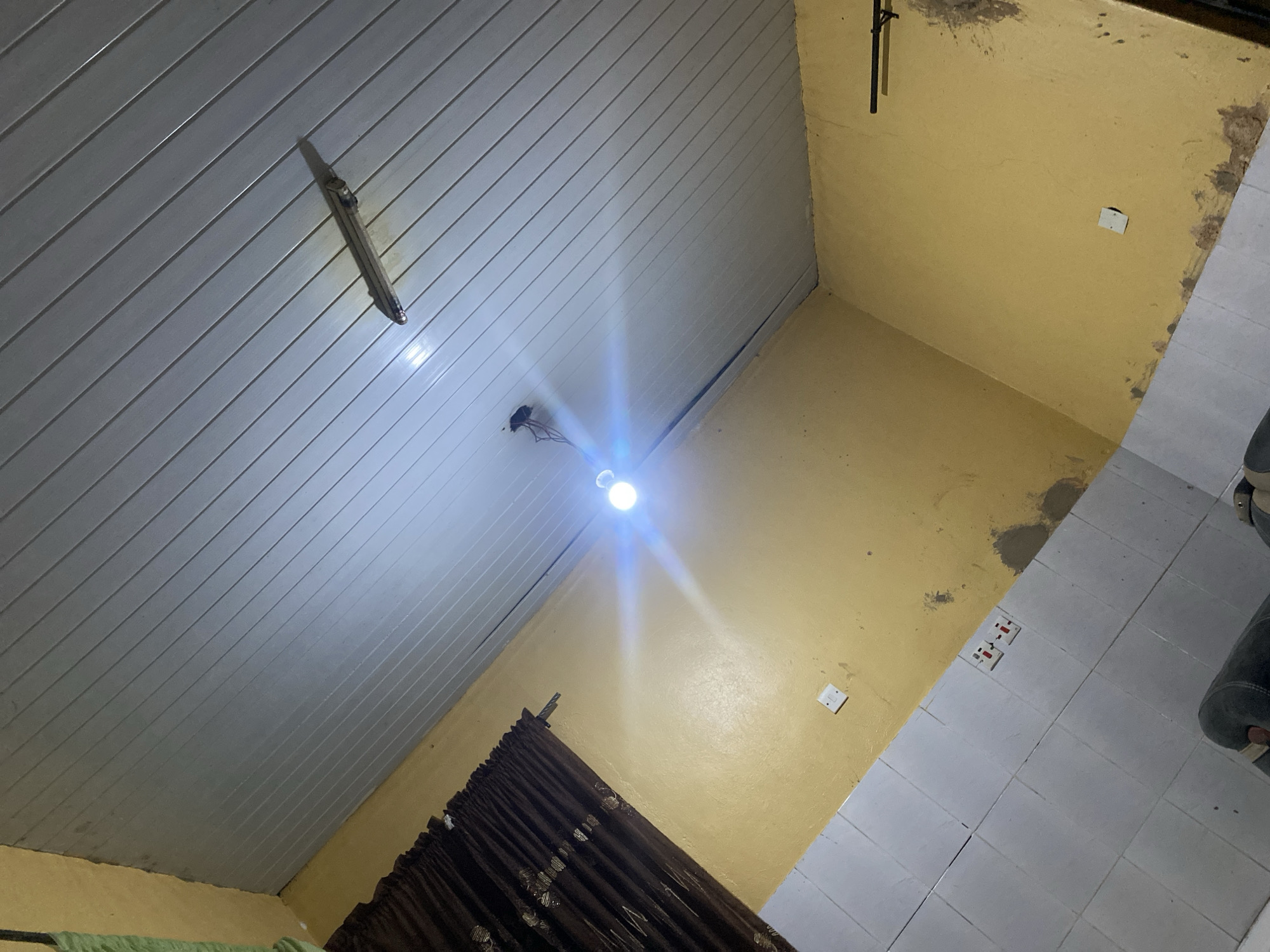 The image presents an indoor room viewed from a low angle, looking upwards towards the ceiling and across the walls and floor. The ceiling is clad in light grey, horizontally paneled material, likely PVC, which reflects the bright illumination. A single, bare light bulb, emitting a strong white light and glare, hangs from the center of the ceiling, serving as the primary light source. Another older, unlit light fixture or housing is attached to the ceiling towards the upper left.
The walls are painted a mustard yellow, showing signs of wear, including patches of discoloration, dampness, or exposed plaster, particularly along the ceiling line and corners. On the right-hand wall, a small white electrical switch or outlet plate is visible. Further down, near the floor, a white double electrical outlet is mounted on the yellow wall. A dark, vertical pipe or conduit runs along the upper right wall.
The floor is partially visible, segmented into white square tiles on the right side and a yellow-painted surface on the left, consistent with the wall color. A dark brown, patterned curtain is drawn across a window or opening, situated along the bottom-center of the frame. Parts of a dark, possibly upholstered object and a green fabric item are visible on the floor at the far right and bottom left, respectively. The strong artificial lighting suggests the scene takes place at night or in a poorly lit environment. This functional room, located in Shen, Nigeria, appears to have modest finishes and some signs of age. No visible text is present.