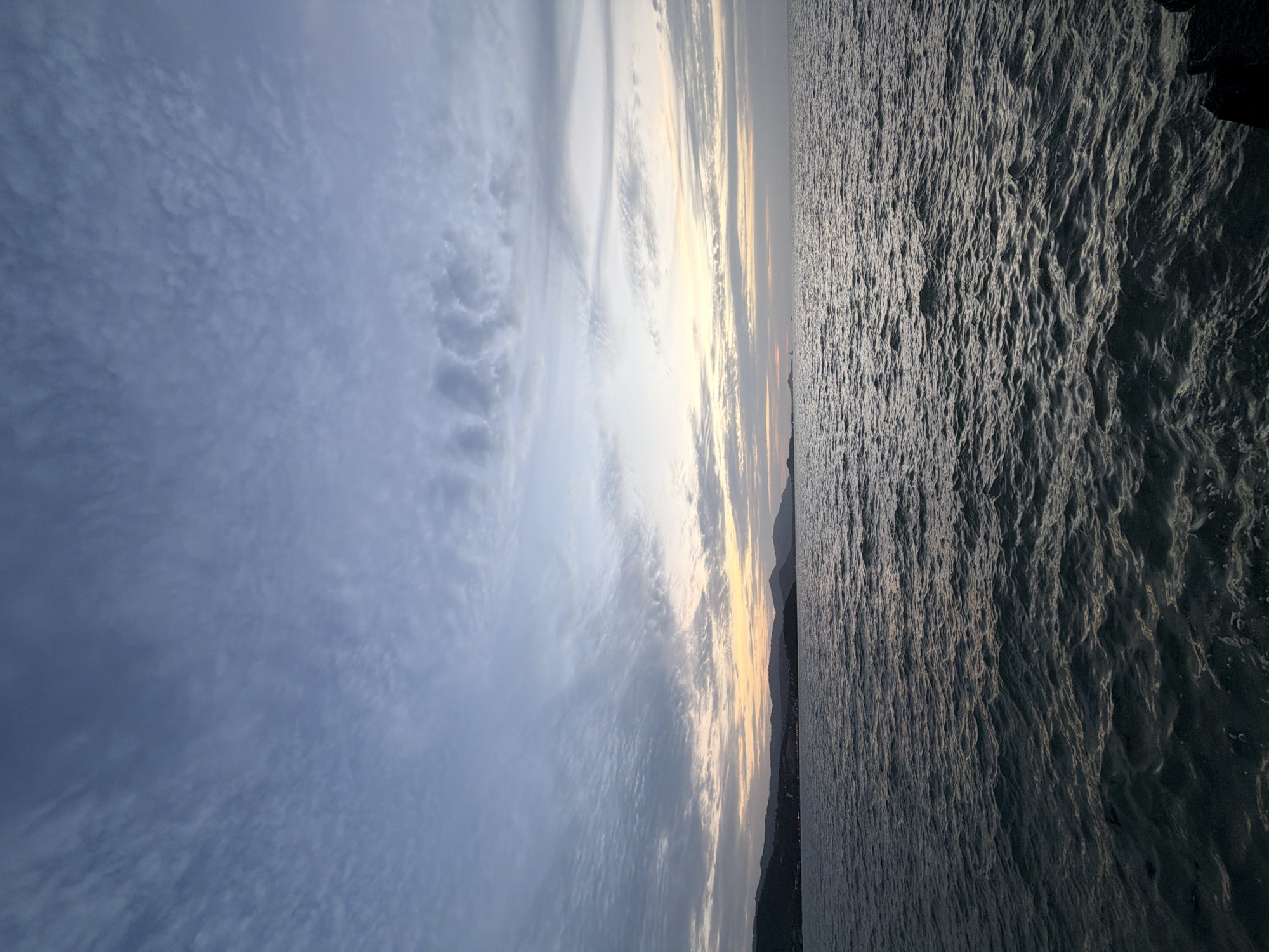 The image shows a seascape at dusk, likely in Ko Samui District, Thailand. The sky is filled with a mix of wispy and textured clouds, with soft hues of orange and yellow near the horizon, indicating the last light of sunset or the first light of dawn. Below the sky, a gentle body of water stretches across the frame, exhibiting small ripples and waves that catch the diffused light. In the distance, a dark, silhouetted coastline with rolling hills is visible, suggesting a landmass bordering the sea. The overall atmosphere is tranquil and serene, characteristic of a coastal view during twilight. There are no people or specific man-made structures prominently featured, emphasizing the natural elements of the scene.