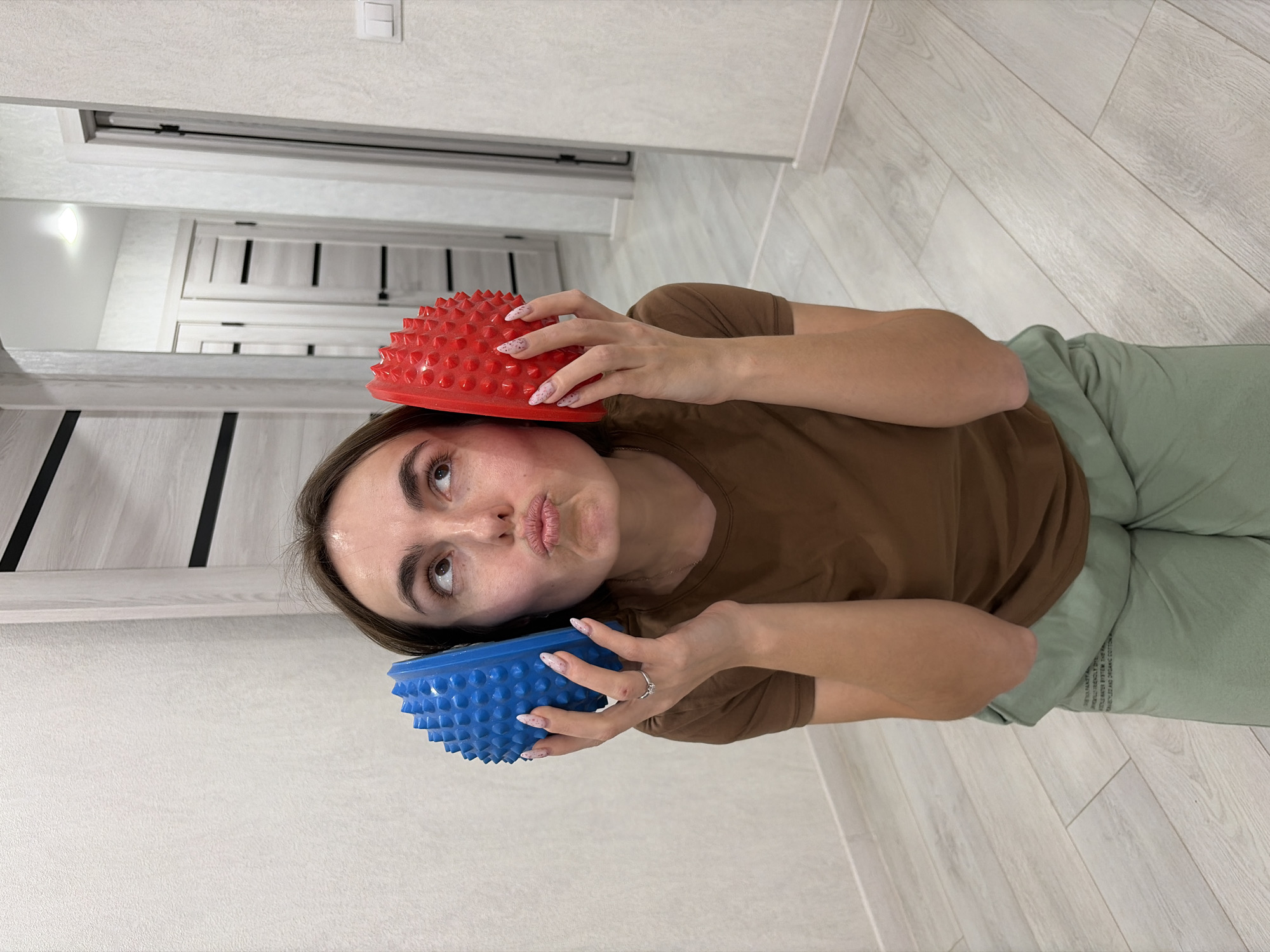 A woman is lying on a light-colored, wood-patterned floor, holding a red, spiky semi-sphere above her head and a blue, spiky semi-sphere under her chin. She is wearing a brown t-shirt and light green pants. The location is inside a room with white walls, a white door with black accents, and a light switch visible on the wall. This scene is likely located in an interior setting, but there is no information to confirm that this location is in the city of Sarapul, Russia.